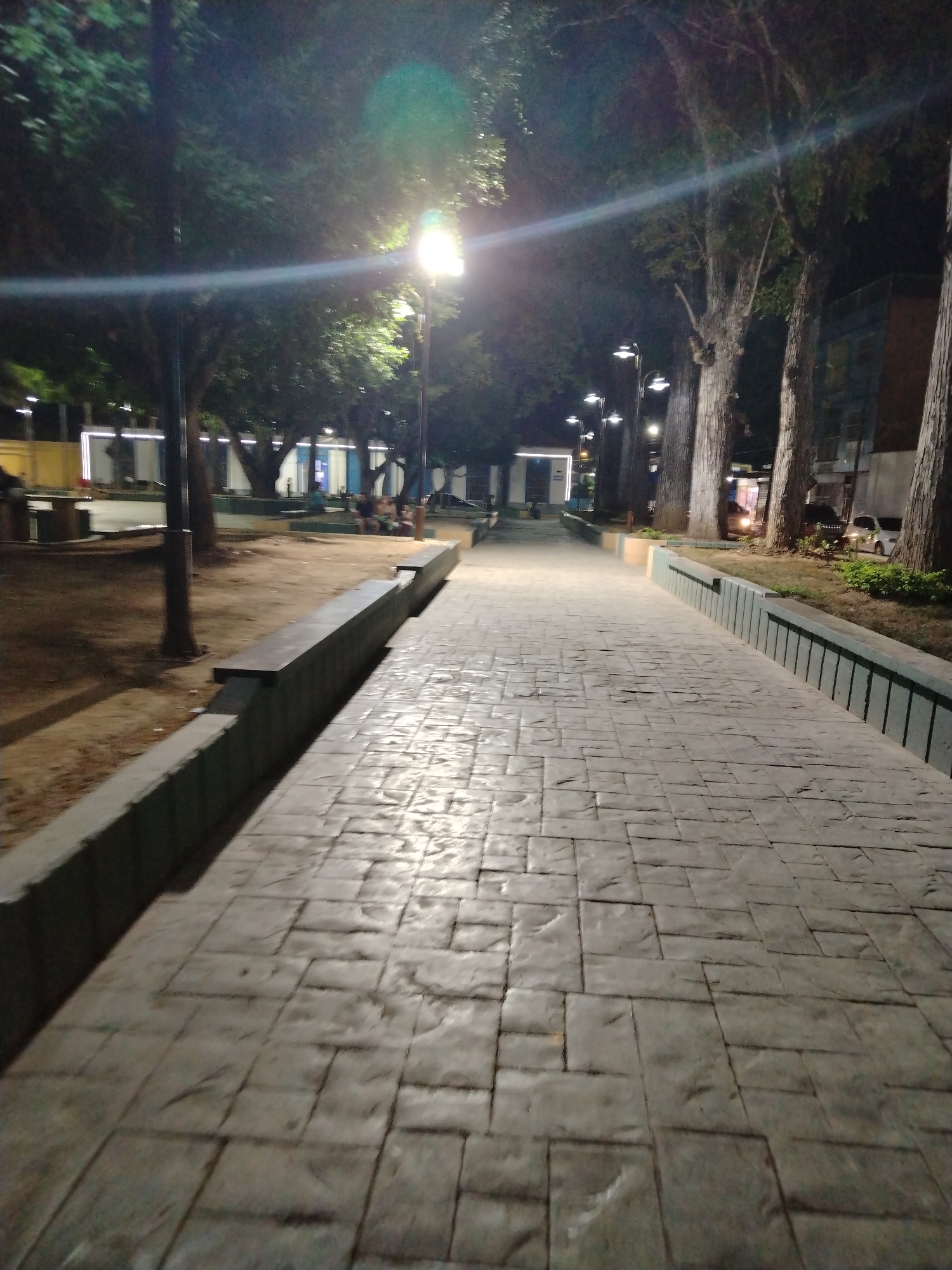 The image shows a nighttime view of a paved walkway in a park or plaza. It is illuminated by streetlights. The walkway extends into the distance, flanked by raised curbs and trees. In the background, there are people on benches. The scene is Tinaquillo, Venezuela, and includes details like the paving stones of the walkway, the design of the streetlights, the benches, and the surrounding buildings and landscaping.