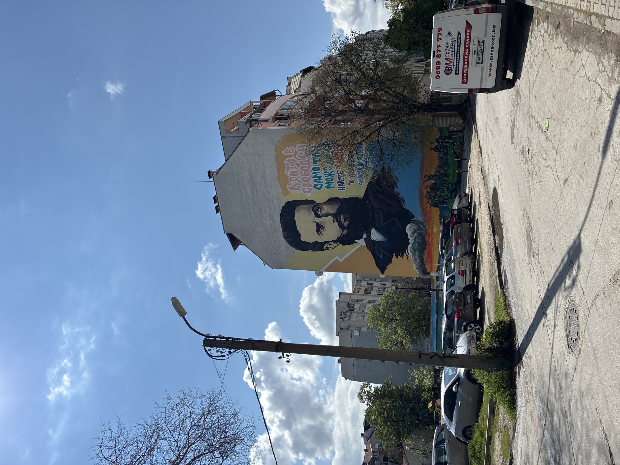 The image depicts a mural of Hristo Botev, a Bulgarian poet and revolutionary, painted on the side of a building. The mural includes a portrait of Botev and text in Bulgarian. To the right of the mural is a white van with a phone number, website, and the phrase "отпушване на канали", which translates to "drainage of canals" in English. Along the side of the street are parked cars. Above the mural, are other buildings and the sky. The photo was taken in Plovdiv, Bulgaria.