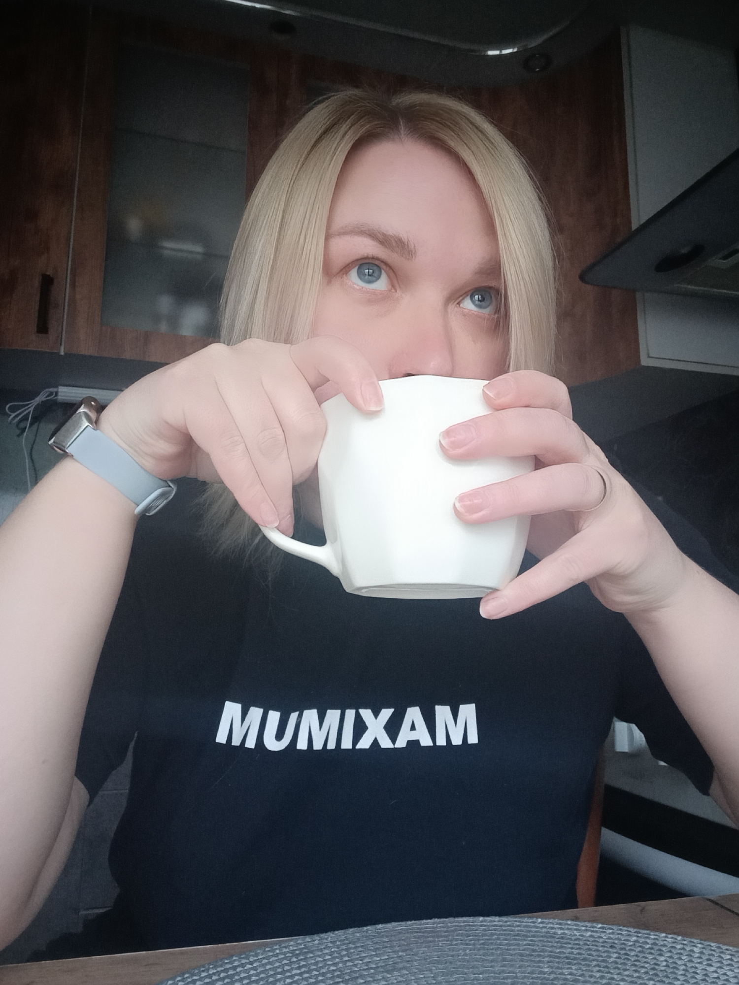 A woman with blonde hair and blue eyes is holding a white mug, likely drinking from it, in what appears to be a kitchen setting. She is wearing a black t-shirt with the word "MUMIXAM" printed in white capital letters. On her left wrist, she has a smartwatch with a gray band. She also has a ring on one of the fingers of her right hand. The background shows kitchen cabinets and appliances, suggesting an indoor setting. There is no indication of the city of Chaikovsky, Russia.