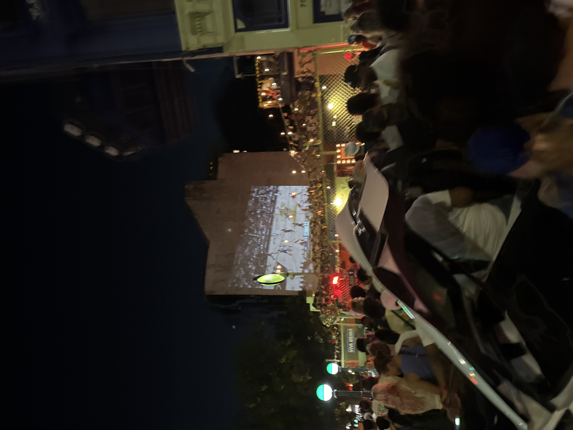 This image captures a vibrant night scene in an urban setting, identified as New York City, United States of America, by the partially visible "718" area code on a building and the overall streetscape.
The scene is densely populated with a large, diverse crowd of people of varying ages and ethnicities, casually dressed, standing, mingling, and observing various points of interest. In the immediate foreground, the white hood and windshield of a car are prominently visible, possibly with its hood slightly ajar.
In the midground, a festive atmosphere is created by numerous string lights and decorations, including a visible sign that appears to read "VIVA MEXICANA" and a smaller "BEER" sign. A fence-like structure, also adorned with lights and small banners, separates the foreground crowd from a raised platform or balcony, which is also populated by people.
A central focal point is a large video projection on the light-colored facade of a building in the background, displaying what appears to be a basketball game or a similar sporting event. Near the projection, a tall pole emits a bright green light. Additional green-glowing streetlights punctuate the left side of the scene. Buildings with distinct architectural features frame the edges of the image, and a dark sky is visible above. The overall environment suggests a lively outdoor gathering or event.