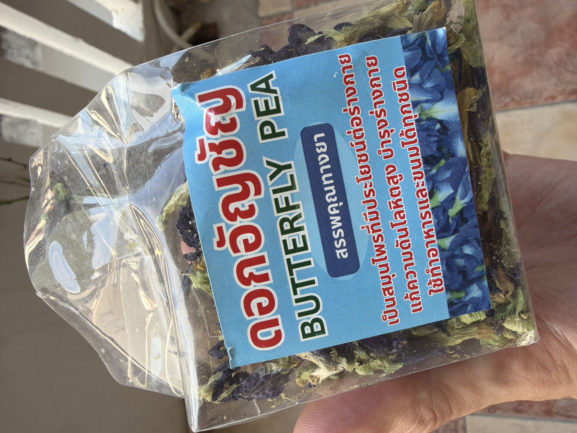 A clear plastic bag filled with dried butterfly pea flowers is held in a person's left hand. A blue label with red and green text is affixed to the front of the bag. The text on the label is in Thai and English. The English text reads "BUTTERFLY PEA". The bag is being held in an outdoor setting, with a tiled surface visible below and a blurry white structure in the background, suggesting it is a balcony or outdoor area. The lighting indicates it is daytime. The location of Budva, Montenegro is not ascertainable from the image.
