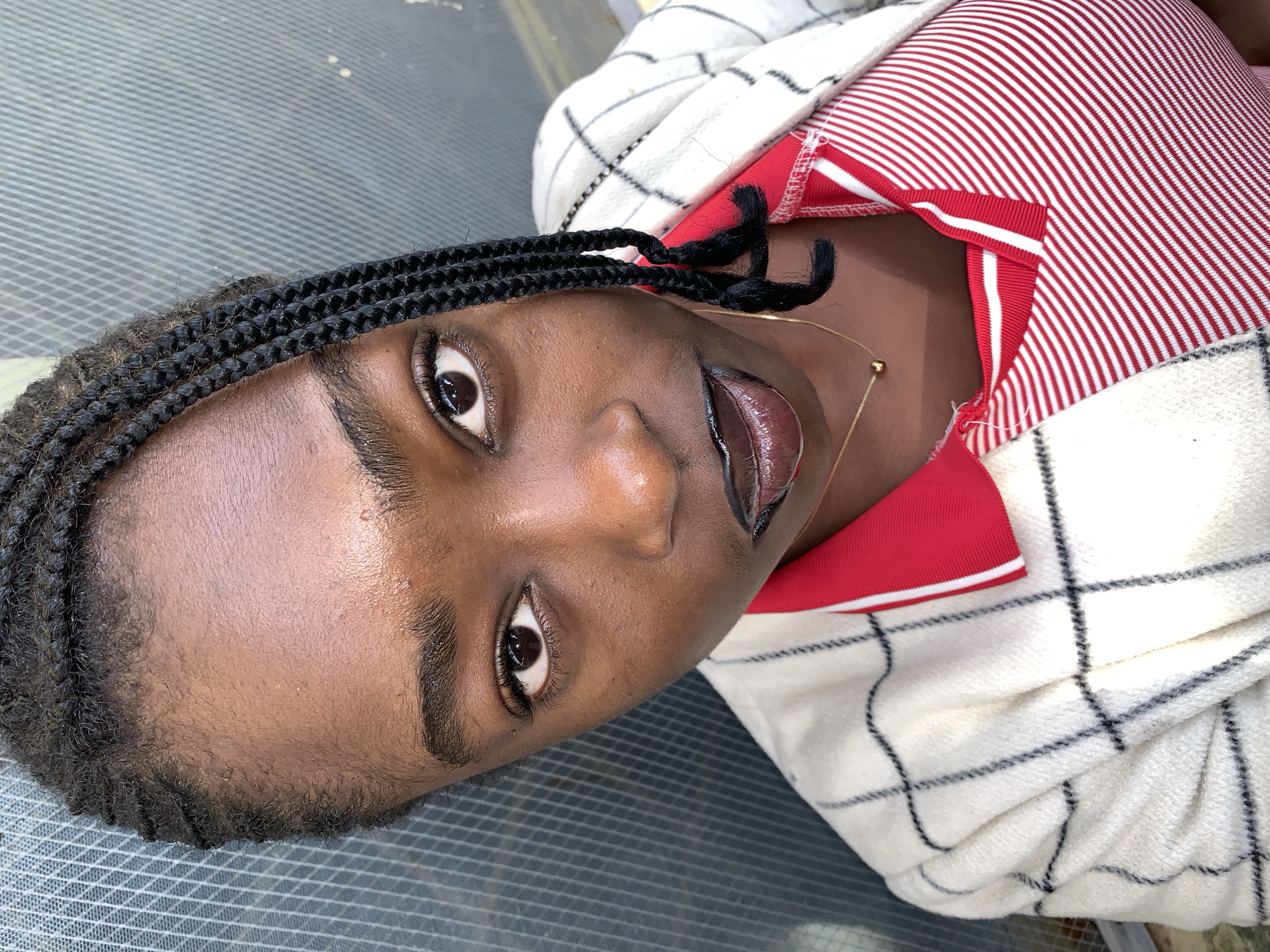 A close-up, overhead shot features a young Black woman with braided hair and dark lipstick. She is wearing a red and white striped polo shirt underneath a cream-colored jacket with a black plaid pattern. The woman is looking directly at the camera, with her face slightly angled. The background is a gray, textured surface with a faint grid pattern, suggesting an outdoor urban setting. The lighting is bright, creating highlights on her skin. The overall impression is a confident portrait.
