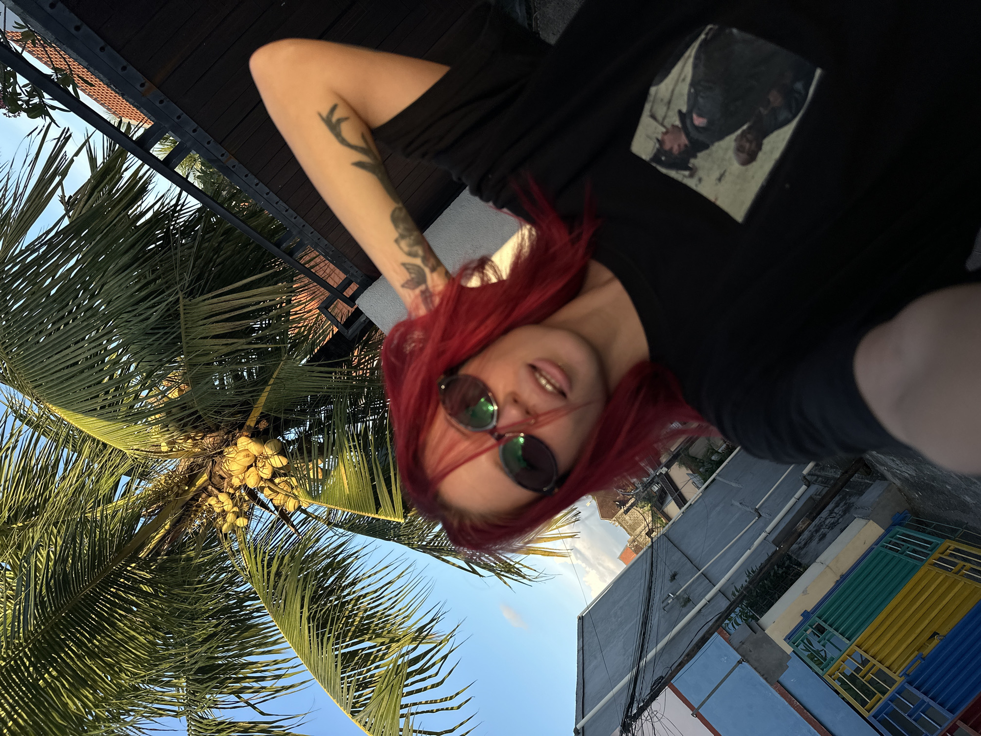 A photograph taken in North Kuta, Indonesia, shows a young person with red hair lying down, wearing sunglasses and a black T-shirt with an image on it, likely a selfie. The shot is taken from a low angle looking up. A palm tree with coconuts is visible to the left, with a clear blue sky in the background. In the opposite direction, there are buildings, with one featuring a colorful (blue, yellow, green) corrugated metal fence. The photo includes power lines running between buildings and tattoos on the visible arm.
