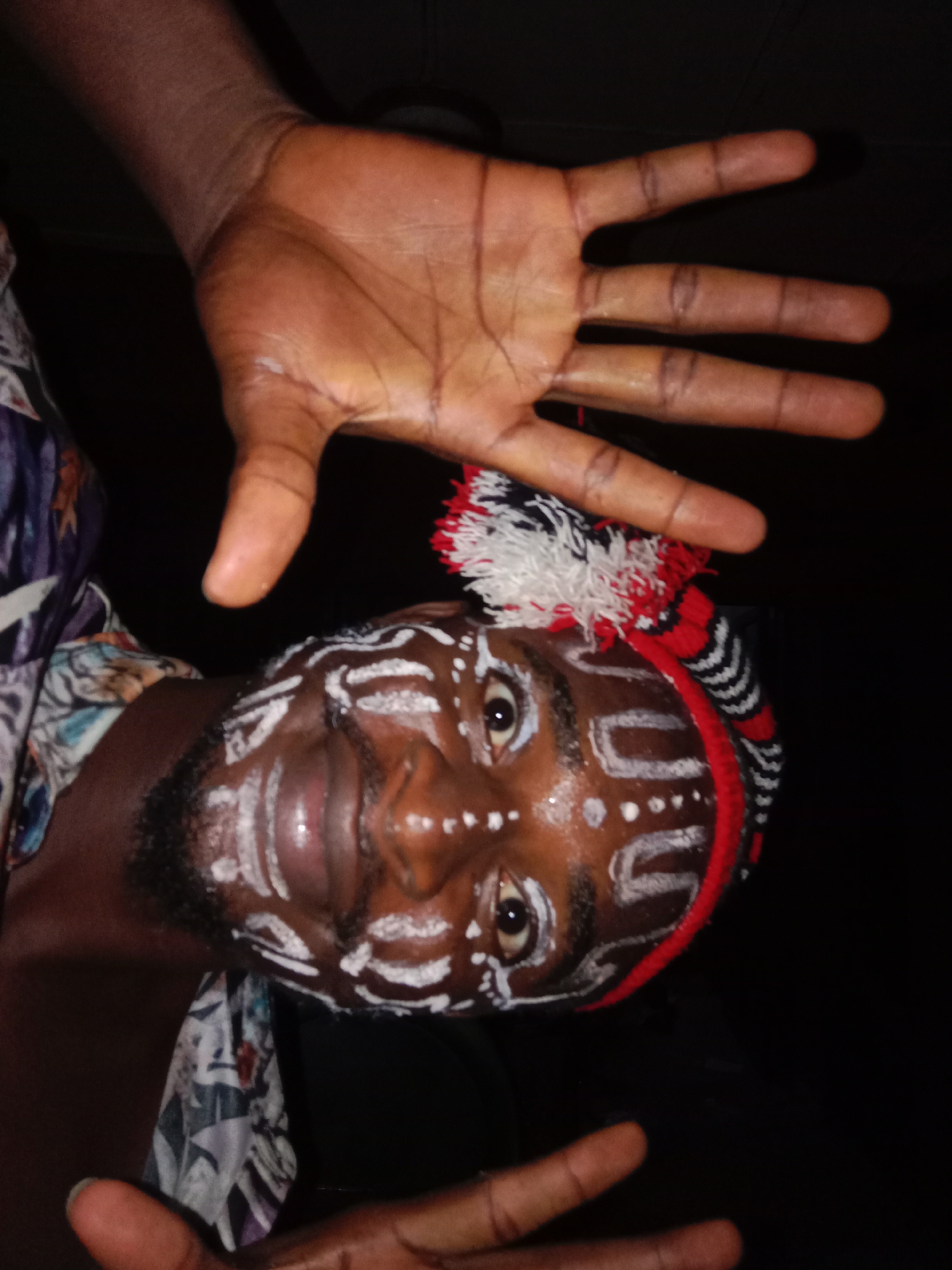 A rotated, close-up portrait features a person with dark skin against a stark, dark background. The subject's face is expressively adorned with white body paint, forming patterns of dots, lines, and U-shapes across the forehead, cheeks, nose, and chin. They wear a distinctive headpiece, likely a knitted cap or headband, with red, white, and black stripes and matching tassels positioned near the top of the head. The person's eyes are open and gaze towards the camera, accompanied by a subtle, engaging smile. One hand is prominently raised above the head, palm facing outwards with fingers spread. A portion of another hand is visible at the bottom left of the frame. To the left of the subject, a patterned garment, possibly a shirt or draped fabric, with hues of blue, purple, white, and black, is visible. The scene is dimly lit, with a light source illuminating the face and the raised hand, creating highlights that accentuate the details of the paint and skin. The dark, featureless background suggests an indoor setting or a night-time event. The overall presentation implies a cultural performance, ritual, or artistic expression. No text is visible.