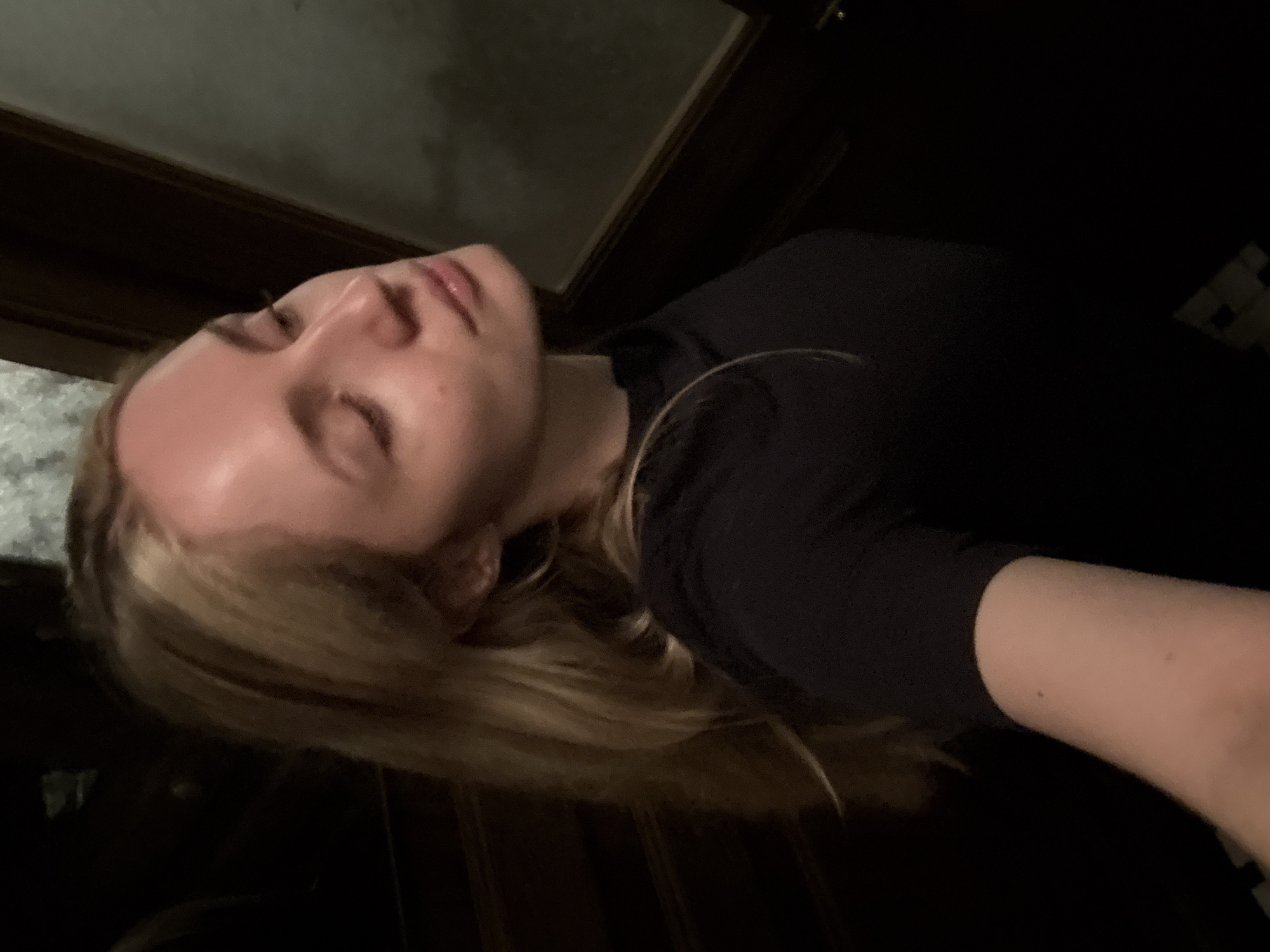 A fair-skinned person with blond hair is lying down. The person is wearing a black shirt. The background is dark and out of focus, but a window is visible. The location is not discernible from the image.