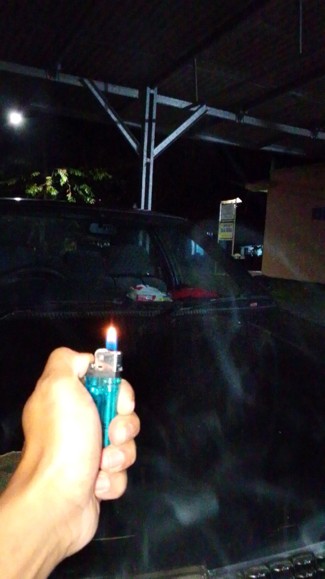 An outdoor, night-time, eye-level shot captures a hand holding a lit blue lighter in front of a dark-colored car. The car's windshield is visible, with what appears to be some items placed on the dashboard. In the background, a building and some trees can be seen. The scene is set under a structure with metal supports, possibly a carport or shelter. The overall lighting is dim, with the flame of the lighter providing a significant light source. The location is identified as Cintaratu, Indonesia.