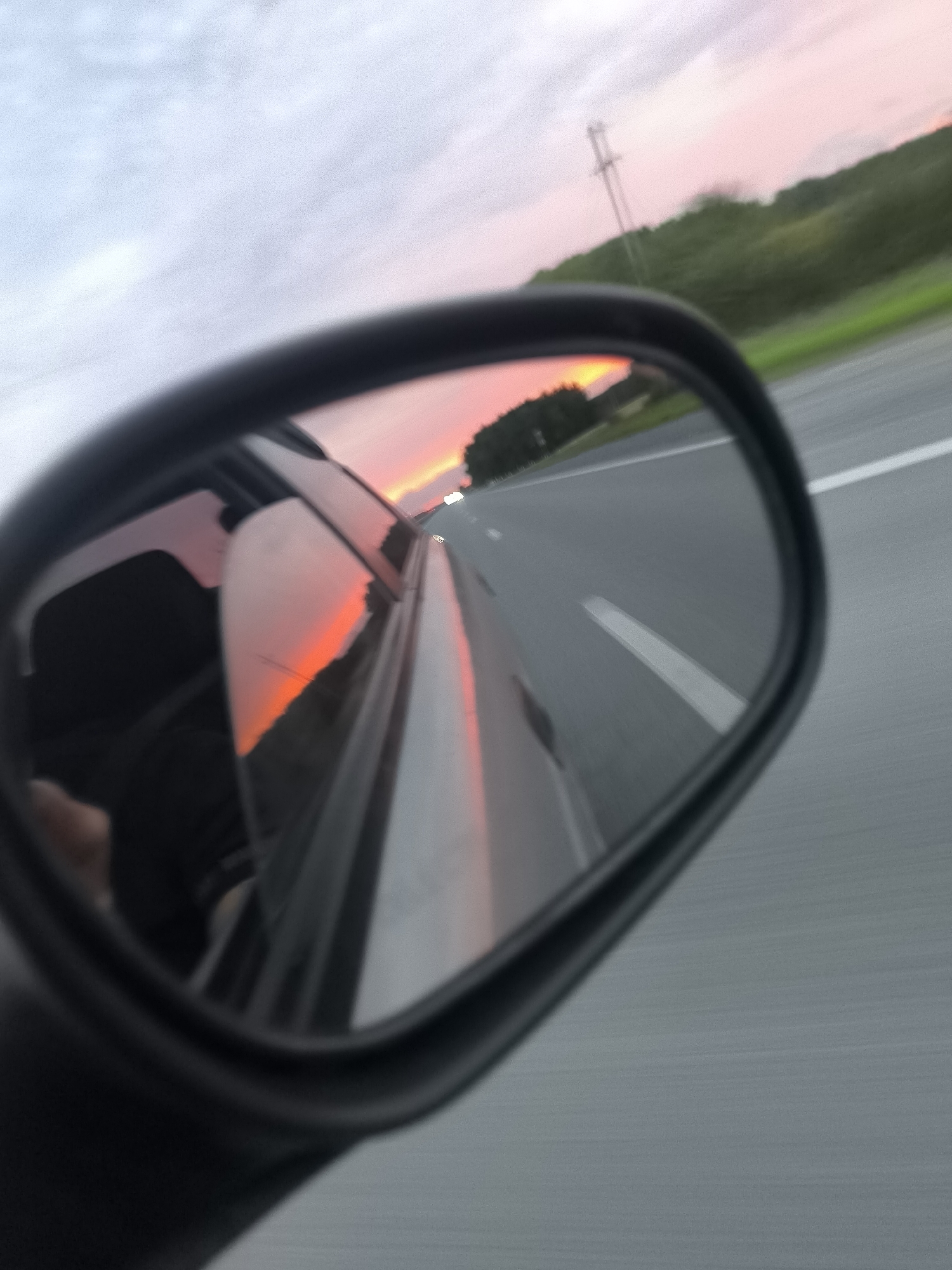 The image shows a side-view mirror of a car, reflecting a sunset with pink and orange hues. The mirror also shows the interior of the car, including a person's arm and shoulder, and the side of the car. The background outside the mirror depicts a highway with green trees and a power line. The scene suggests a road trip or commute during sunset. The location of Krasnyi Yar, Russia is not discernible from the image.