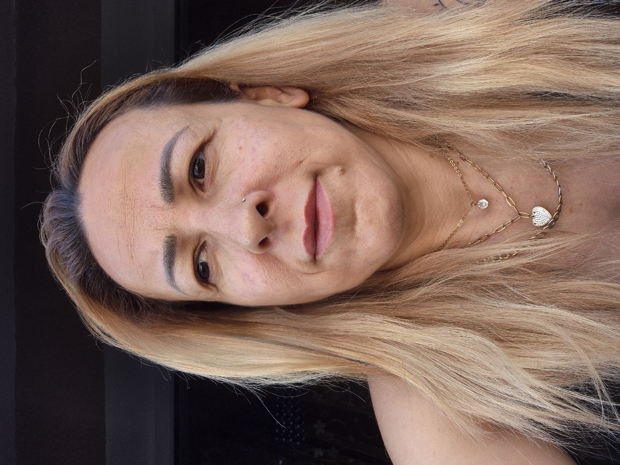 A close-up, head-and-shoulders portrait shows a light-skinned person with long, blonde hair that is pulled back slightly from their face. They have brown eyes, dark eyebrows, and a small nose stud. The person is looking to the left of the frame. Two gold necklaces are visible: one with a small, clear pendant and another with a heart-shaped pendant adorned with small white stones. The background is dark and out of focus. The lighting suggests an outdoor setting, possibly during the day, with the person facing a light source. The person's skin shows some texture and faint wrinkles.