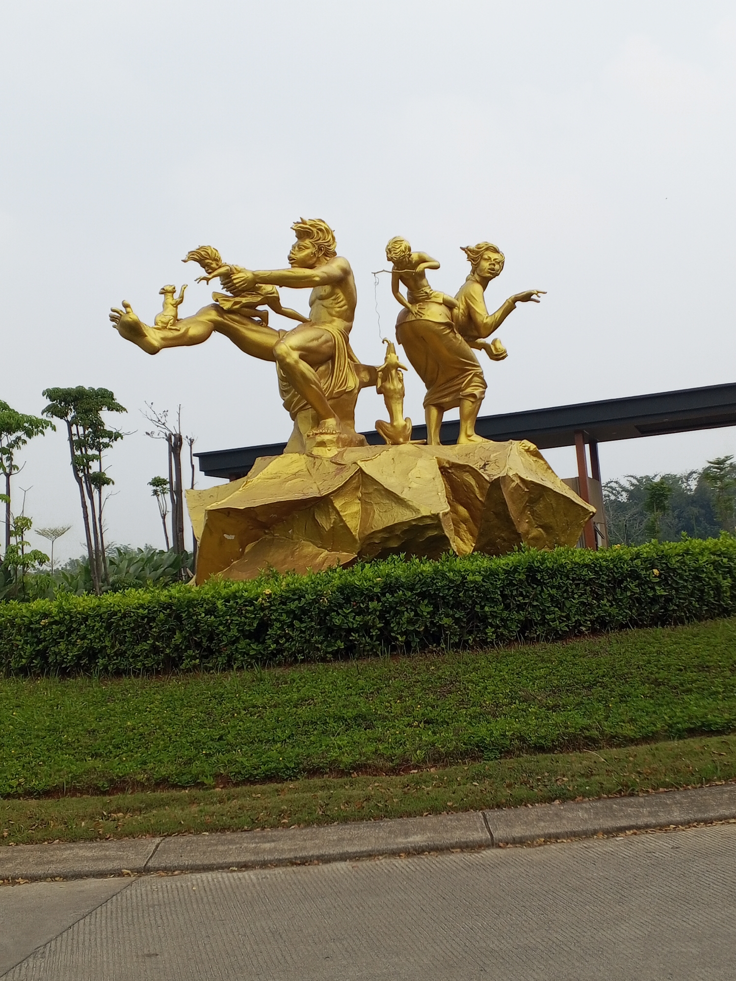 A golden sculpture depicting two adults and two children, along with a dog, is situated on a rocky pedestal. The sculpture is located in Serang, Indonesia, and is surrounded by manicured green bushes and grass. In the background, trees and a building with a dark roof are visible. The sky is overcast. The scene appears to be a public space or park.