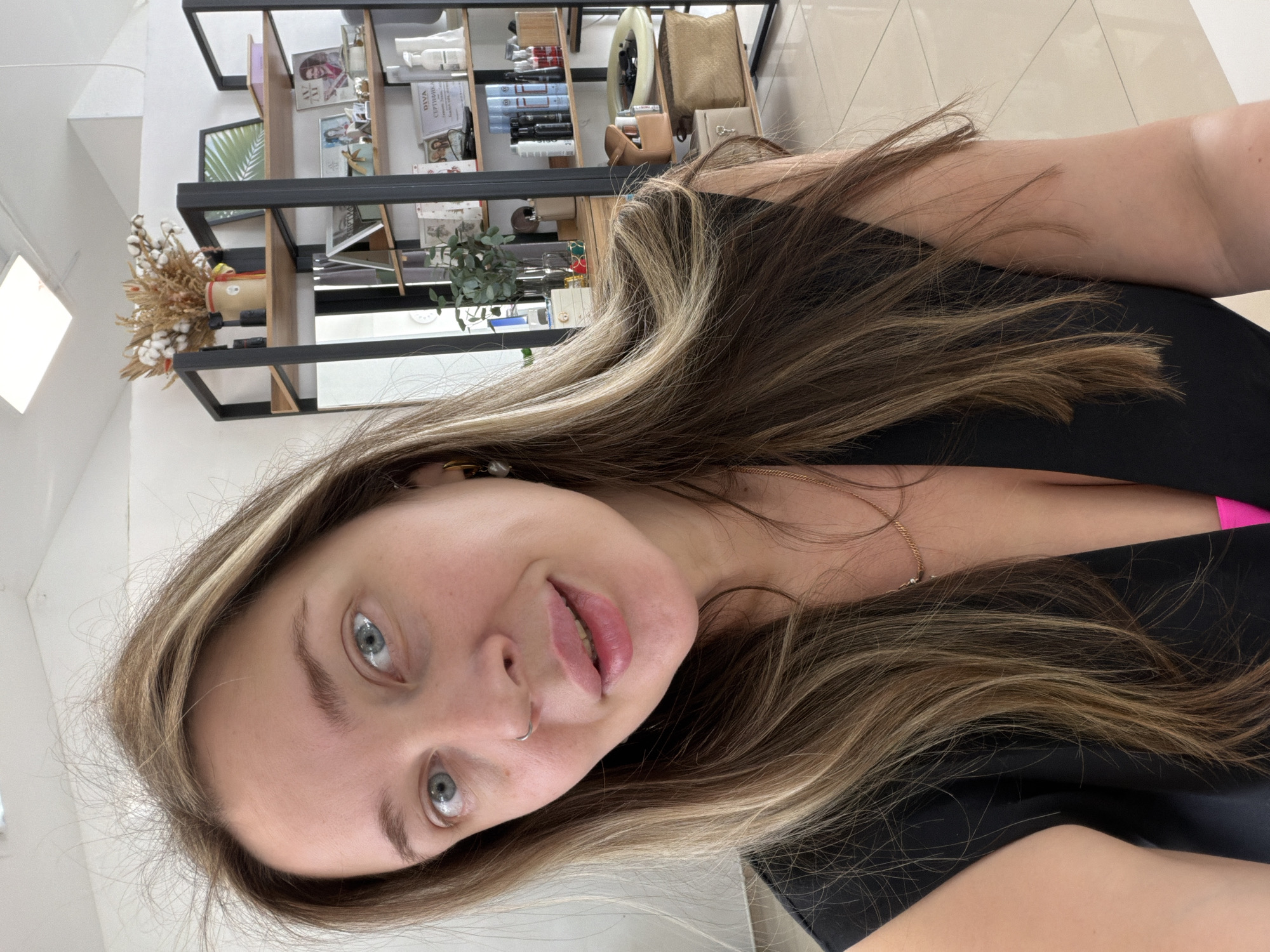 A close-up selfie depicts a young woman with light skin, gray-blue eyes, and light brown hair with blonde highlights. She is wearing a black top. A gold chain necklace is visible around her neck. She has a silver nose ring in her left nostril. The background reveals the interior of a room, where shelves display various cosmetic and hair care products. There is also a glimpse of decorative plants and dried flowers on the shelves. This could be a beauty salon or store. The precise location in Chaikovsky, Russia, cannot be determined based solely on this image.