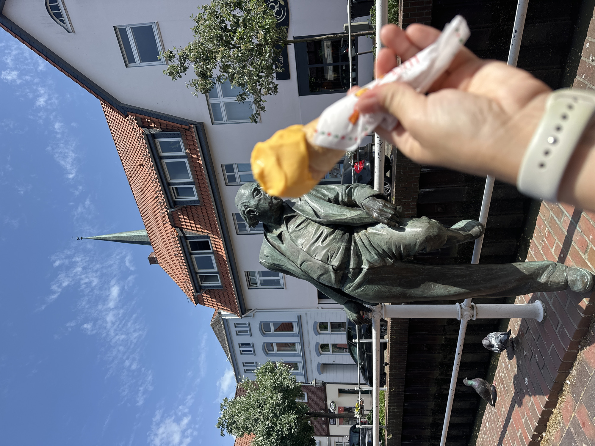 The image depicts a bronze statue of Wilhelm Kieselbach in Buxtehude, Germany. The statue is positioned along the canal, and it appears someone is holding an ice cream cone near the statue's head. The background includes buildings with red tile roofs and white facades with numerous windows. A street with cars is visible behind the statue. Two pigeons are near the statue's feet on a brick surface, and a person's hand wearing a white smartwatch is holding the ice cream. The sky is blue with some clouds.