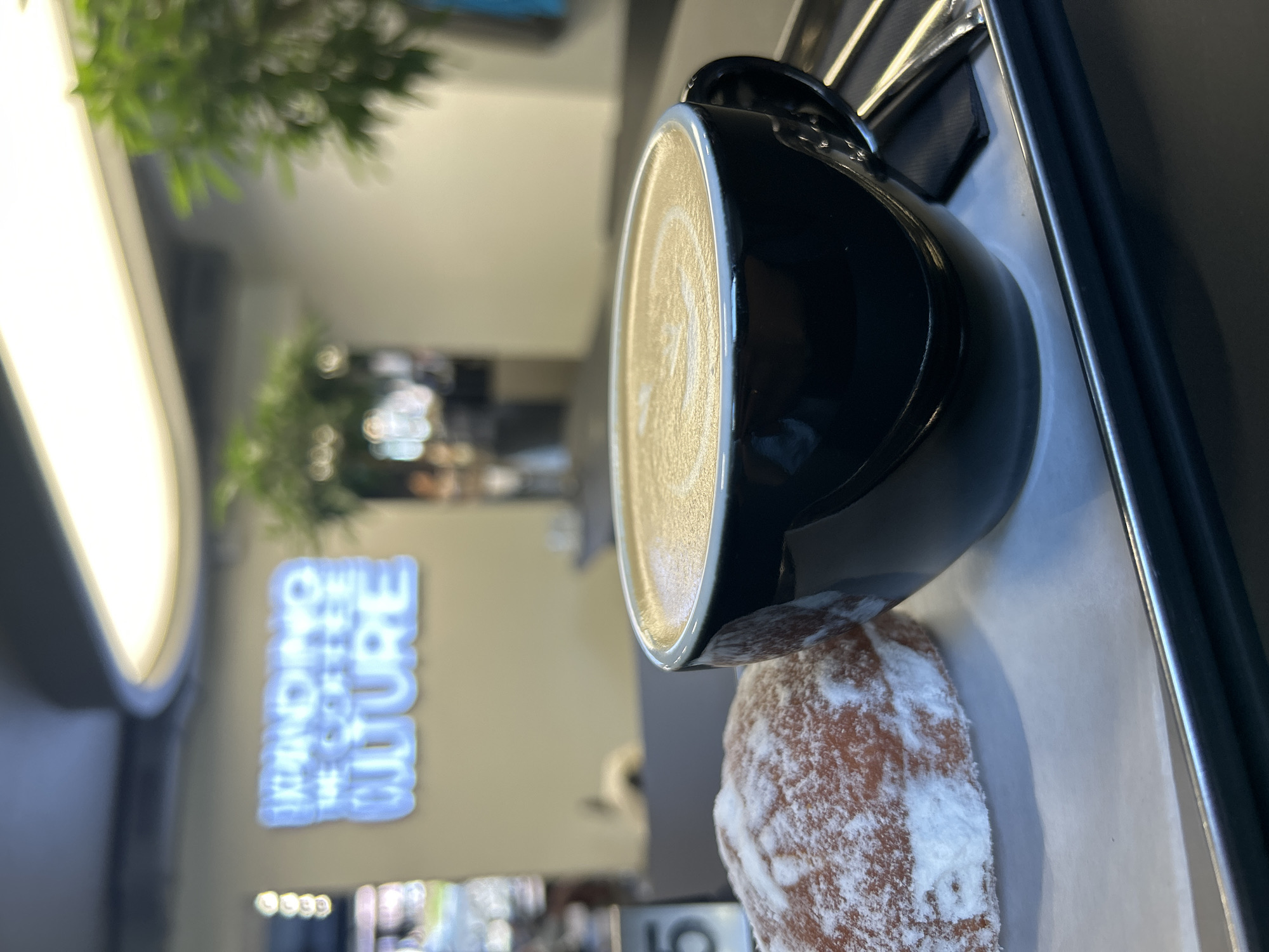 The image shows a black coffee mug with latte art sitting on a white tray with a powdered donut. The background is a blurred interior of a cafe with a sign that reads "INTRODUCING THE COFFEE CULTURE". Based on the sign visible in the photo, the location is likely in Izhevsk, Russia.