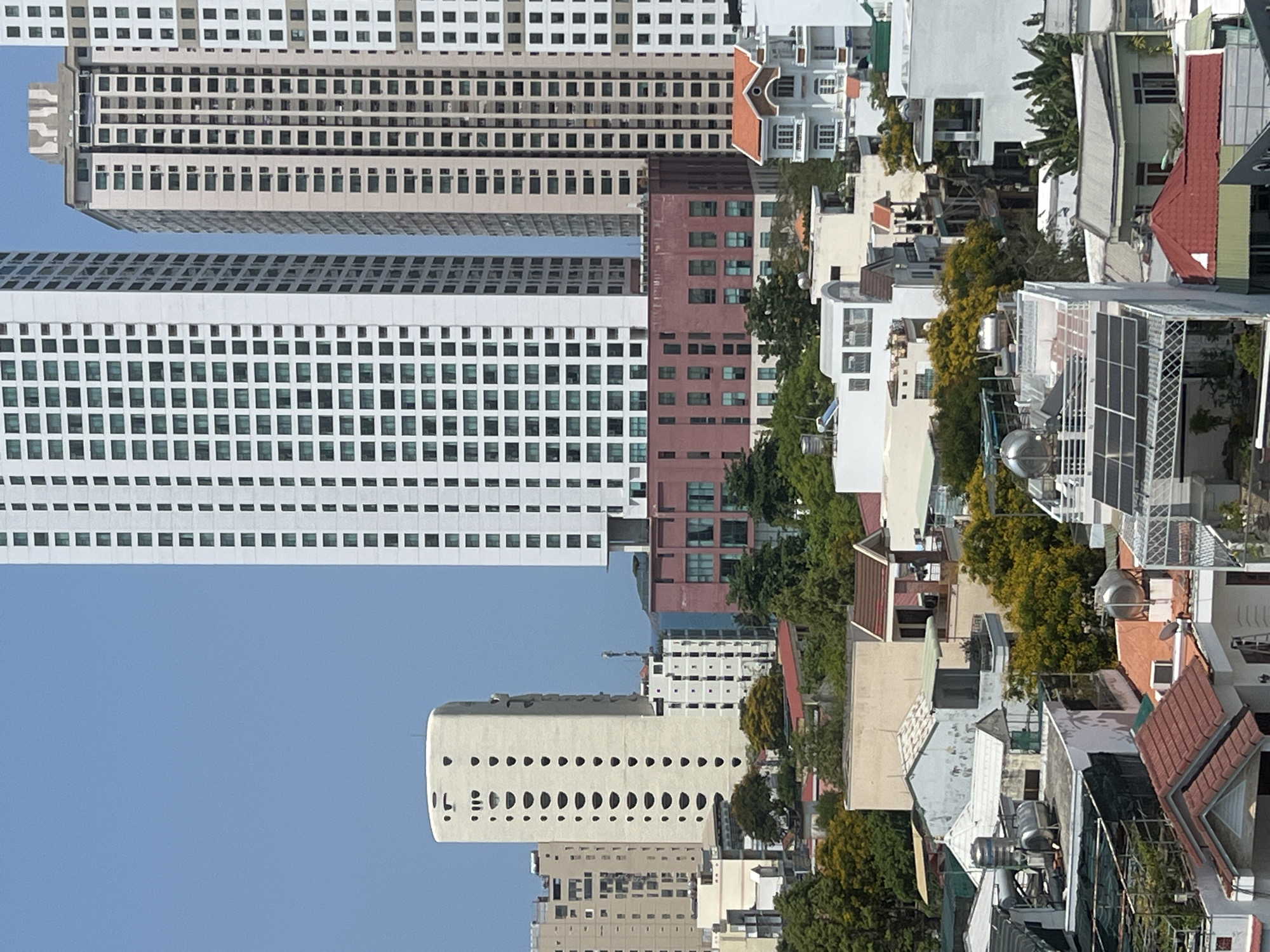 The image presents an urban landscape, likely in Nha Trang, Vietnam, characterized by a mix of high-rise buildings and lower residential structures. The high-rise buildings, some with distinctive architectural features like rows of rounded window openings, dominate the skyline. Their facades are mostly white or light-colored, with uniform patterns of windows suggesting residential or commercial use.
Below, a dense arrangement of smaller buildings, likely residential homes and shops, is visible. These structures exhibit a variety of roof types, materials, and colors, including red tile roofs, corrugated metal, and concrete. Vegetation, including trees and shrubs, is interspersed among the buildings, contributing to the urban greenery.
The sky is clear and blue. The image is a snapshot of a densely populated urban area in Southeast Asia.