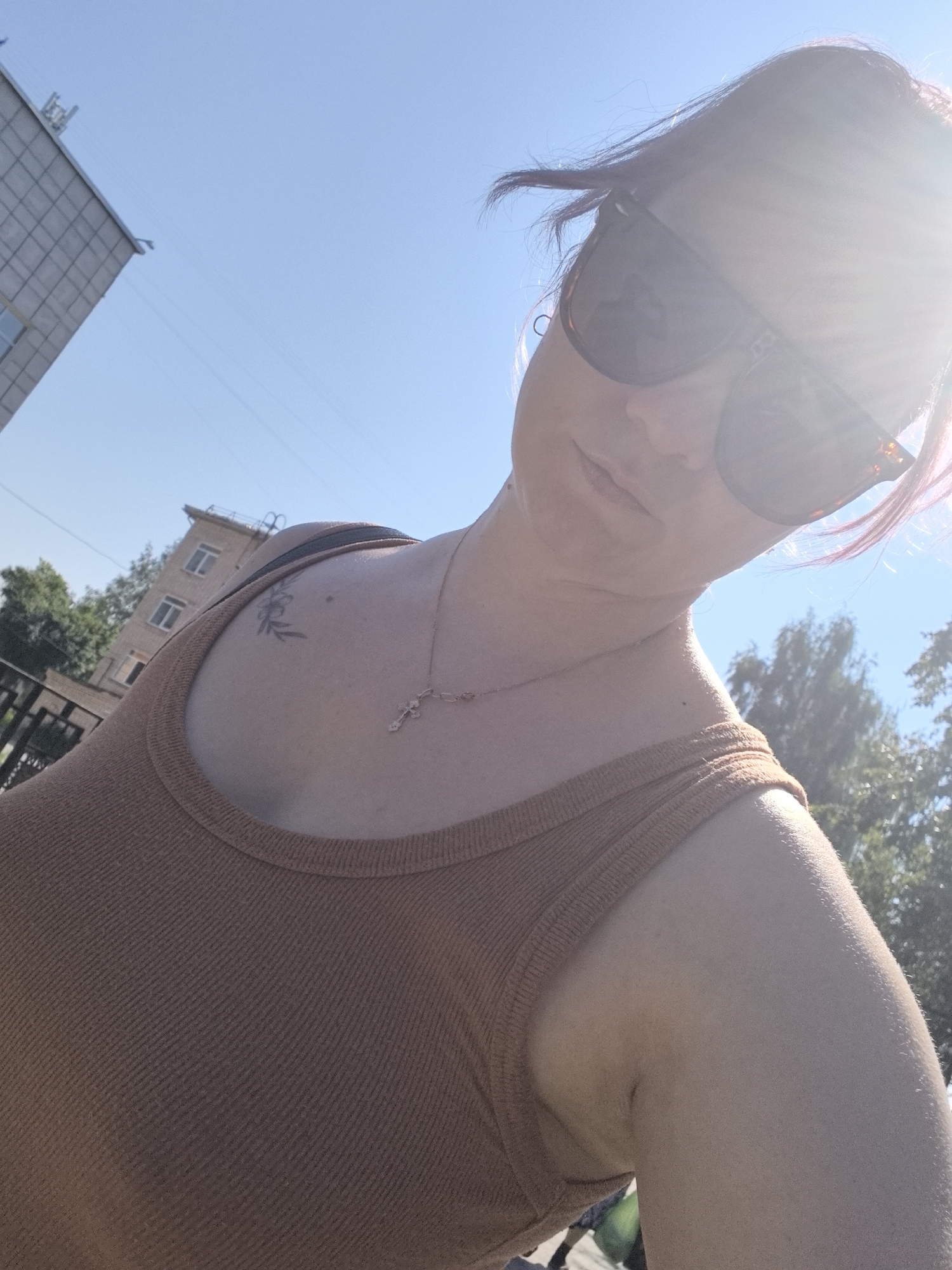A low-angle, wide shot captures a person in Chaikovsky, Russia, on a clear, sunny day. The person, wearing a brown tank top and sunglasses, is looking at the camera with their head tilted. A subtle tattoo of a flower can be seen on their shoulder. In the background, a multi-story building with a gray, tiled facade is visible, along with some trees. The bright sunlight creates a lens flare in the upper right corner of the image.