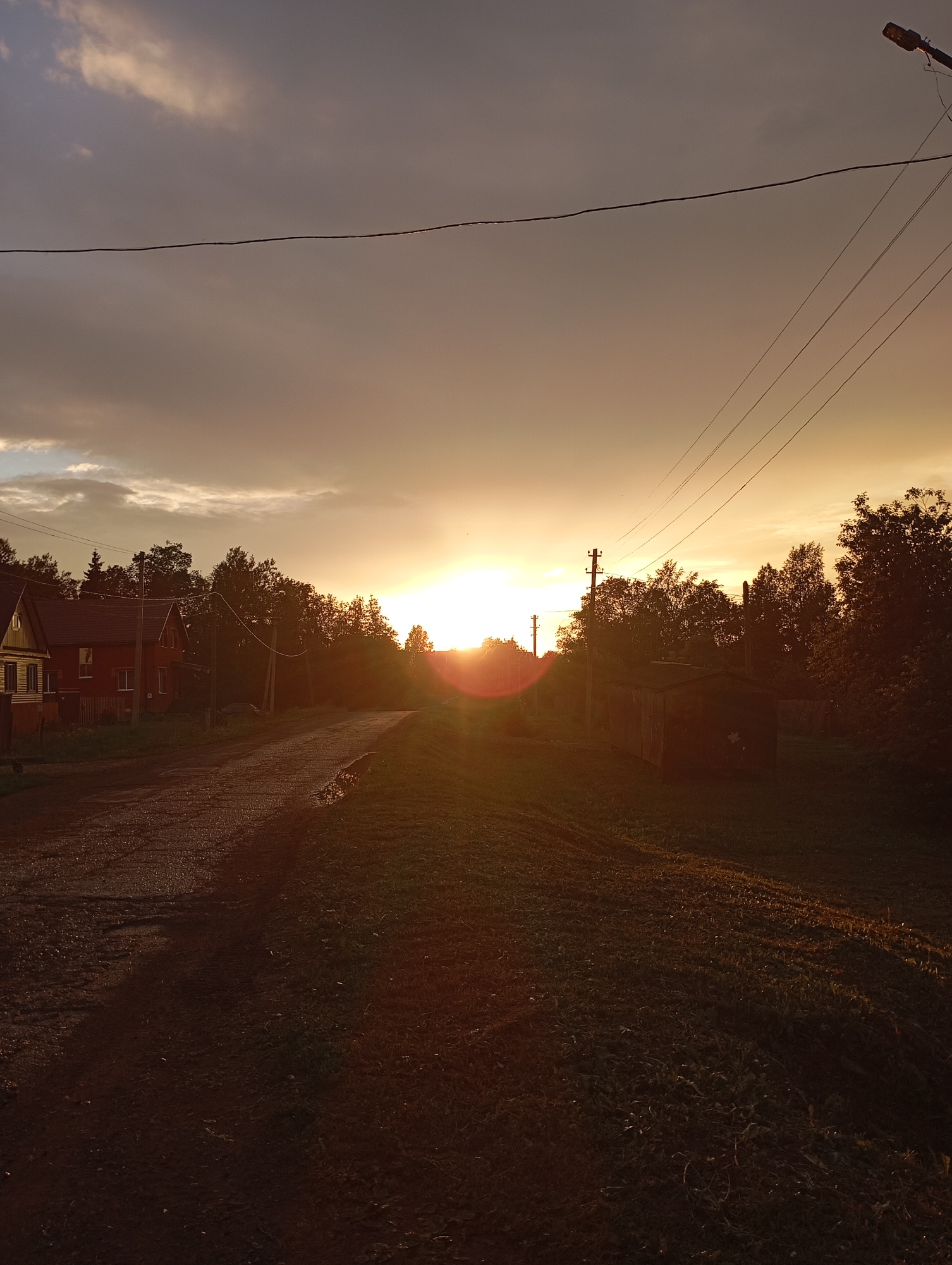 The image depicts a rural sunset scene in Ryabki, Russia, characterized by a dramatic sky transitioning from dark clouds to warm golden hues. The foreground features a gravel road, appearing wet and reflecting the sunset light. To the left, two houses are visible; one is yellow with white trim and a red roof, and the other is red with a darker roof. A wooden fence surrounds the property of the yellow house. Dense trees line the right side of the road, casting long shadows. A small, shed-like structure with a dark roof sits amidst the trees on the right. Power lines crisscross the sky, supported by utility poles, and a street lamp is visible in the upper right corner. The overall atmosphere is tranquil, with the setting sun casting a warm glow over the landscape.