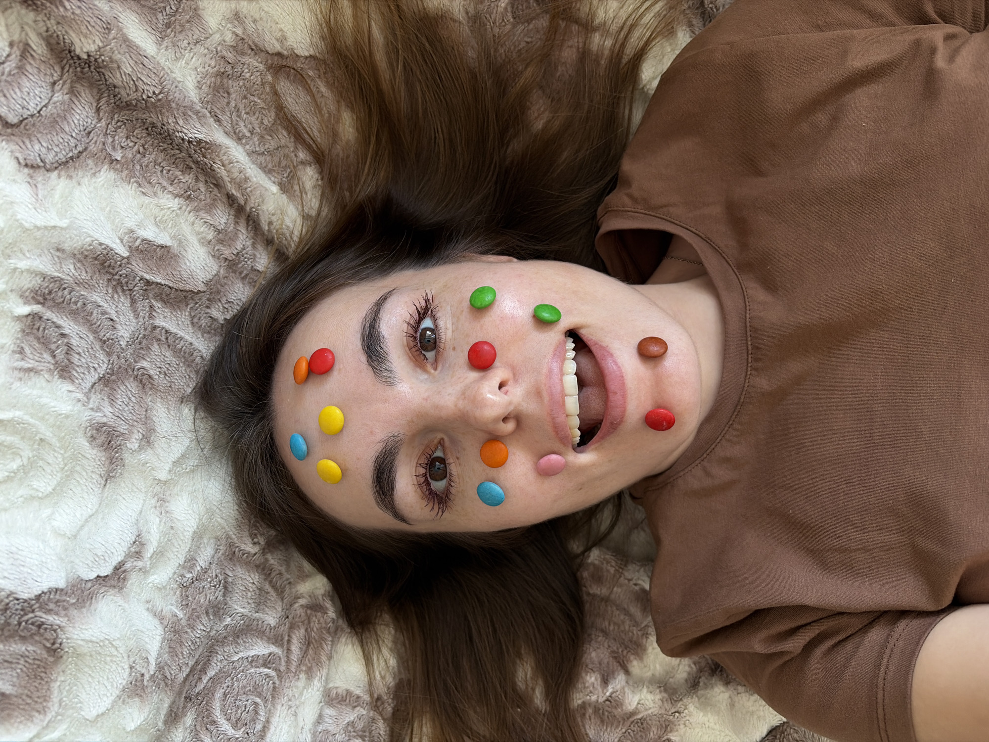 A woman with brown hair and a brown t-shirt is lying on a brown and white patterned blanket. She is facing the camera with an open mouth and colored candy adhered to her face. The background is out of focus. There is no information in the image to indicate it was taken in Sarapul, Russia.