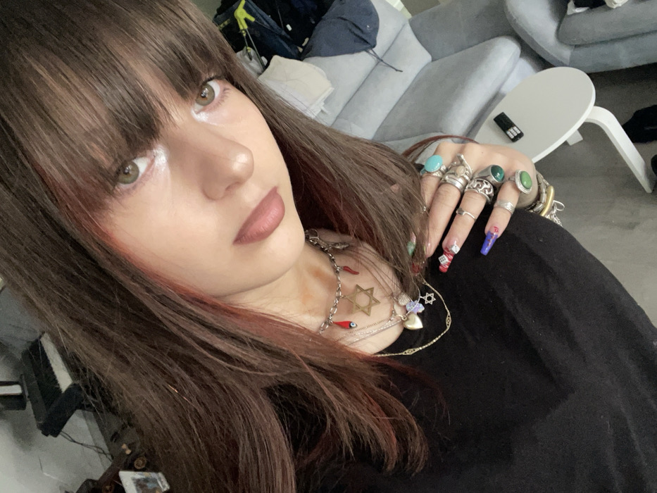 An image taken in an indoor residential setting in Raanana, Israel, depicts a young individual from the chest up, facing the camera at an angle. The individual has long, dark brown hair with reddish highlights and bangs, light-colored eyes, and fair skin with minimal makeup, including light lipstick. They are wearing a black top.
Around the neck, multiple necklaces are visible: a choker-style chain features charms including a brass-colored Star of David, small red chili peppers, and a blue evil eye bead. Layered beneath are thinner chains, one with a small heart charm and another with a smaller Star of David charm.
The individual's left hand is partially visible, featuring long fingernails adorned with intricate nail art in red, white, blue, and checkered patterns, including an ombre effect. Numerous rings, primarily silver-toned with some green and turquoise stones, are worn on multiple fingers.
The background shows a cluttered indoor environment. To the left, a dark-colored electronic keyboard or synthesizer is partially visible on a stand, alongside other indistinct objects including a framed picture. To the right, a light gray upholstered sofa or chair is present, with a white, circular-top side table positioned beside it. A black remote control with silver buttons rests on the side table. Clutter, possibly clothing, is visible on the sofa/chair in the background. The lighting is consistent with an interior space.