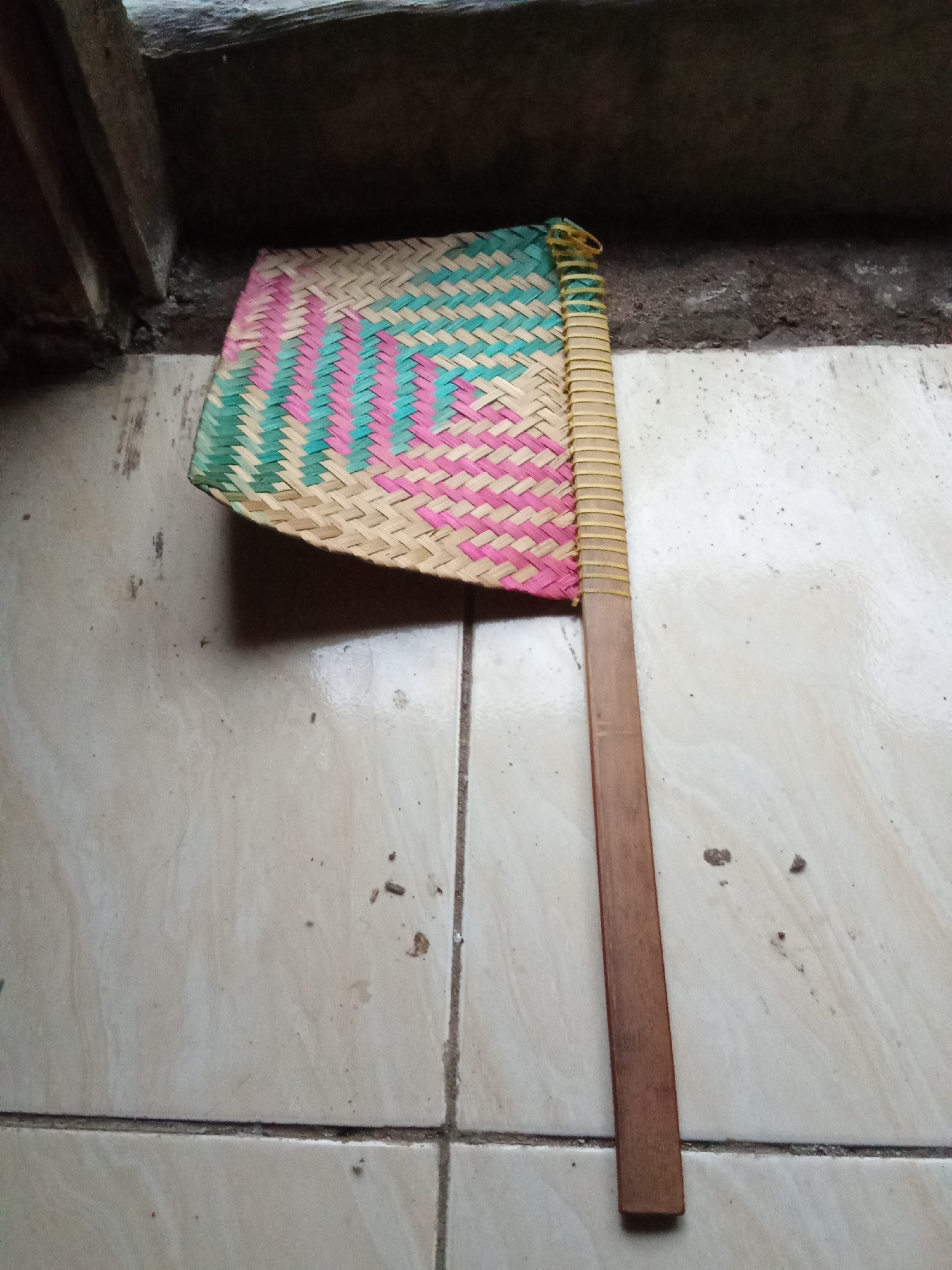 A handheld fan made of woven bamboo with colored strips of pink and teal is lying on a tiled floor. The fan has a light brown wooden handle and is positioned diagonally across the frame, with the fan head on the left and the handle extending towards the bottom right. The tiled floor appears to be made of light-colored ceramic tiles with visible grout lines. In the background, a dark, rough surface, possibly a wall or ledge, is visible. The scene is indoors, likely within a building, and the lighting suggests it is daytime. The image does not contain any people or evidence of human activity other than the fan itself. The location is not specified within the image, but if this were taken in Pemalang, Indonesia, it would be a common household item found in many homes in the region.