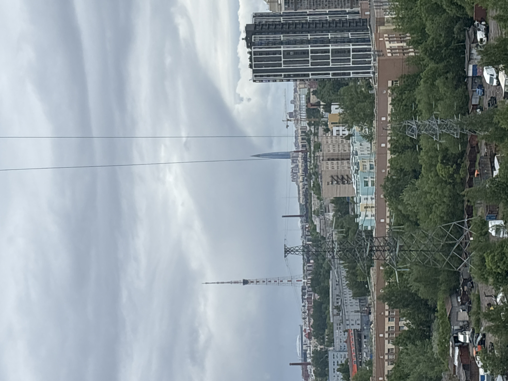 The image shows a cityscape under a cloudy sky. In the distance is a tall spire-shaped building and a red and white broadcast tower. Power lines run across the upper portion of the image. Various buildings and green trees are visible. The setting appears to be an urban area.