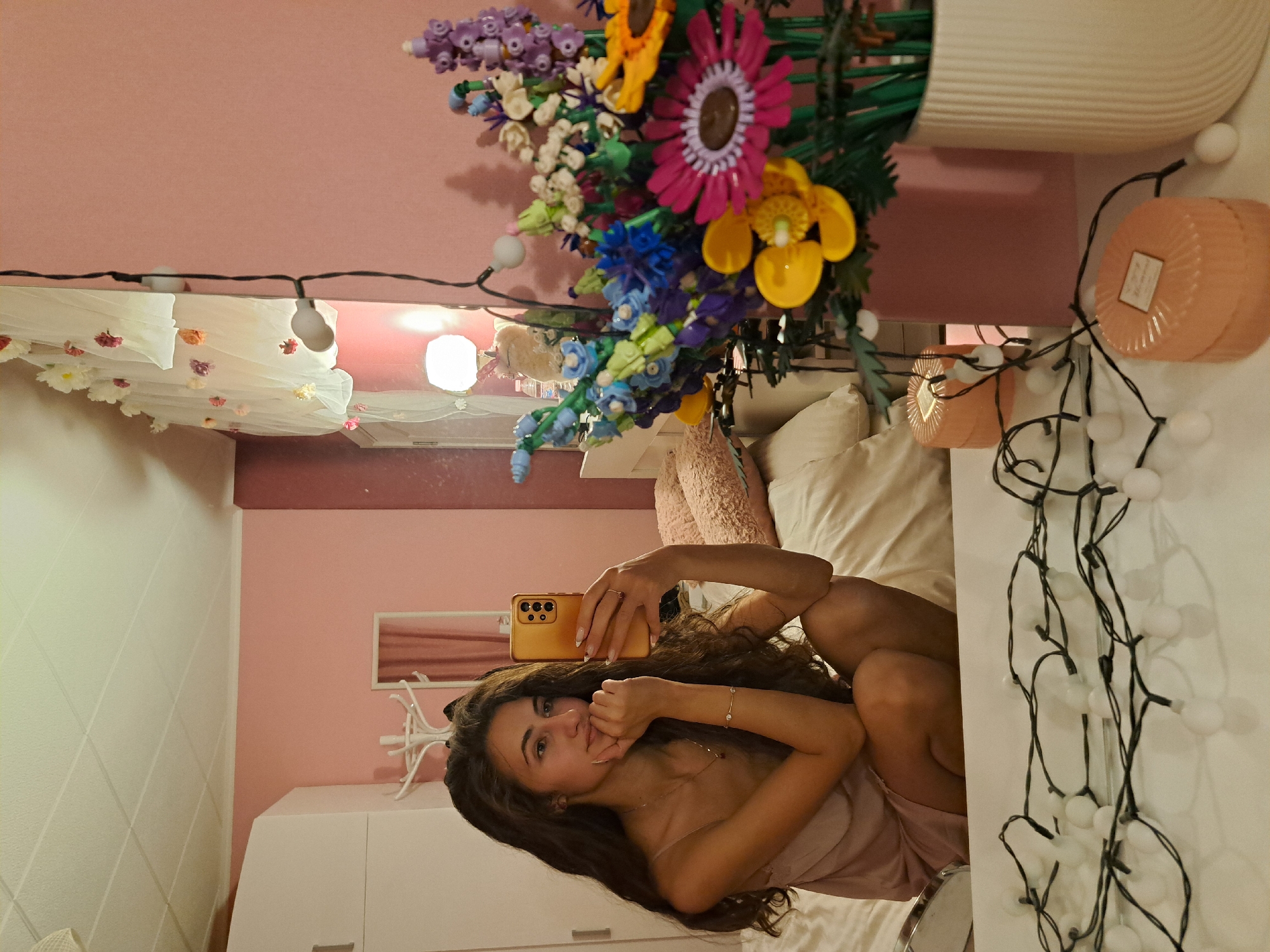A young woman with long, dark, wavy hair is captured in a selfie taken in what appears to be a mirror, or directly with her phone, while she is sitting or reclining indoors. She is wearing a light-colored, possibly pink or beige, strapless garment. Her expression is neutral with a slight smirk, and one hand is raised towards her chin. She holds a gold or orange-colored smartphone with a visible camera array. A ring adorns her finger and a bracelet is on her wrist, while her fingernails are manicured.
The setting is a room, likely a bedroom, characterized by dominant pink walls and a lighter, possibly white, ceiling. One wall features a subtle geometric textured pattern. The room is warmly decorated with various items. Prominently featured is a large bouquet of colorful artificial flowers, resembling Lego pieces, with hues of purple, yellow, pink, blue, white, and green stems, arranged in a white ribbed vase.
String lights with white spherical bulbs are draped throughout the scene, visible above a bed or seating area and along a white surface. Over what appears to be a bed, a sheer white canopy or fabric is adorned with small artificial flowers in shades of red, white, pink, and orange. To the right, two peach or orange cylindrical decorative containers, possibly candles, are visible, one with a label showing the text "HOME." A white hat or coat rack with multiple prongs stands on a white dresser or cabinet in the background. The overall ambiance is cozy and feminine, likely captured in the evening or at night given the use of decorative lights.