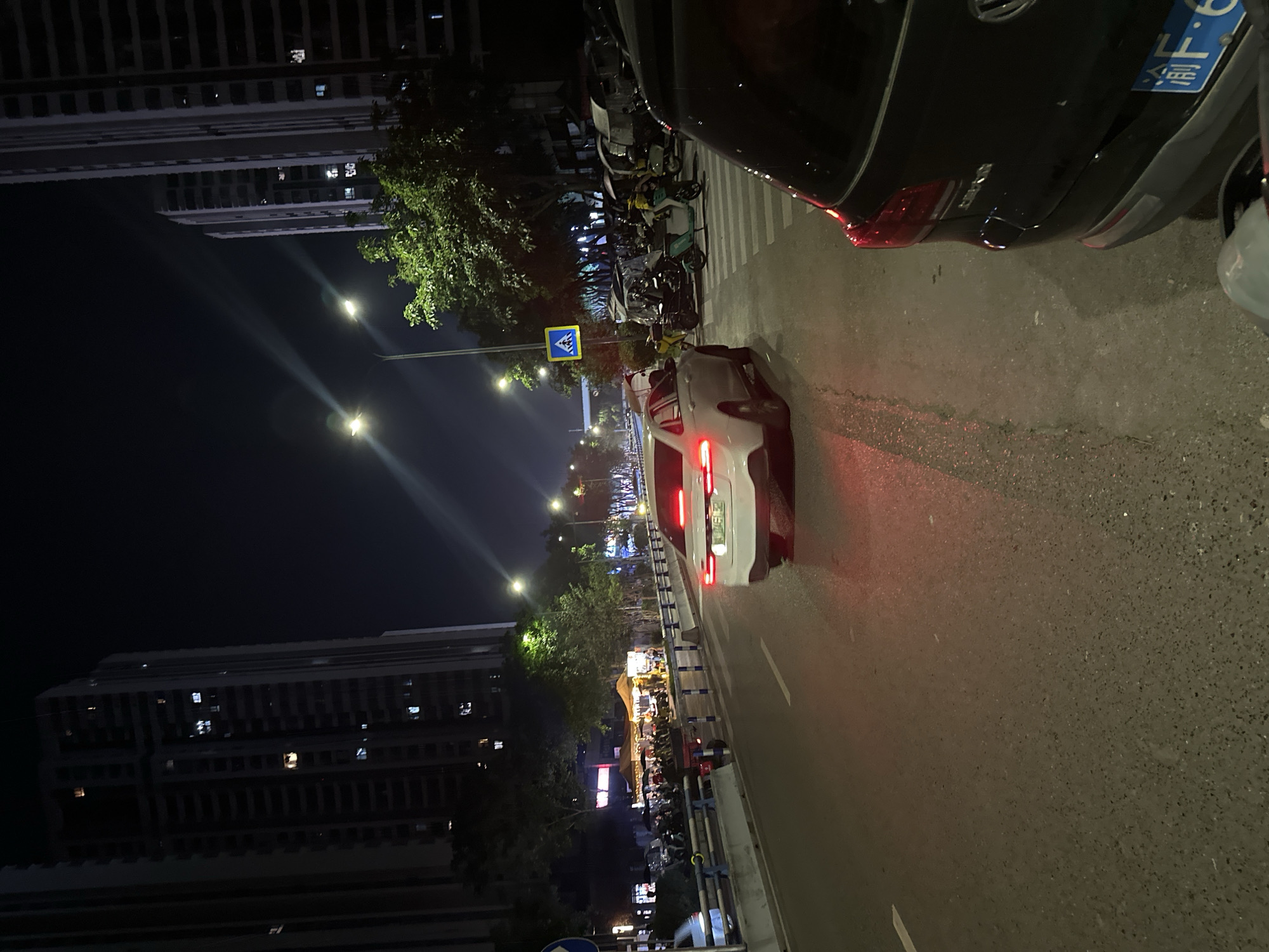The image depicts a nighttime urban street scene in the Chongqing High-tech Zone, China. The viewpoint is from the right side of an asphalt road, looking forward into the street.
In the immediate foreground on the right, the rear quarter of a black SUV is visible, featuring a blue license plate with characters beginning with "渝F". The asphalt road, marked with a white dashed lane line, recedes into the distance. A white sedan with illuminated red taillights is positioned centrally on the road, heading away from the viewer.
To the left of the road, a pedestrian area is separated by blue and white barriers. This area contains multiple illuminated vendor tents, under which several individuals are present, suggesting a street market or food stalls. A row of parked motorcycles and scooters lines this section. Beyond these, green trees are visible, bordering tall, multi-story buildings.
On the right side of the road, beyond a visible pedestrian crossing marked with white stripes, a collection of parked vehicles includes numerous motorcycles, scooters (some appearing to be delivery vehicles), and covered three-wheeled electric vehicles. A blue square sign featuring a white pedestrian crossing symbol on a pole is prominent in this area.
Tall, dark high-rise buildings, consistent with modern urban architecture, are situated on both sides of the street, with various windows illuminated against the dark night sky. Overhead, multiple streetlights emit bright light, and linear light streaks are visible in the upper part of the sky, originating from light sources.