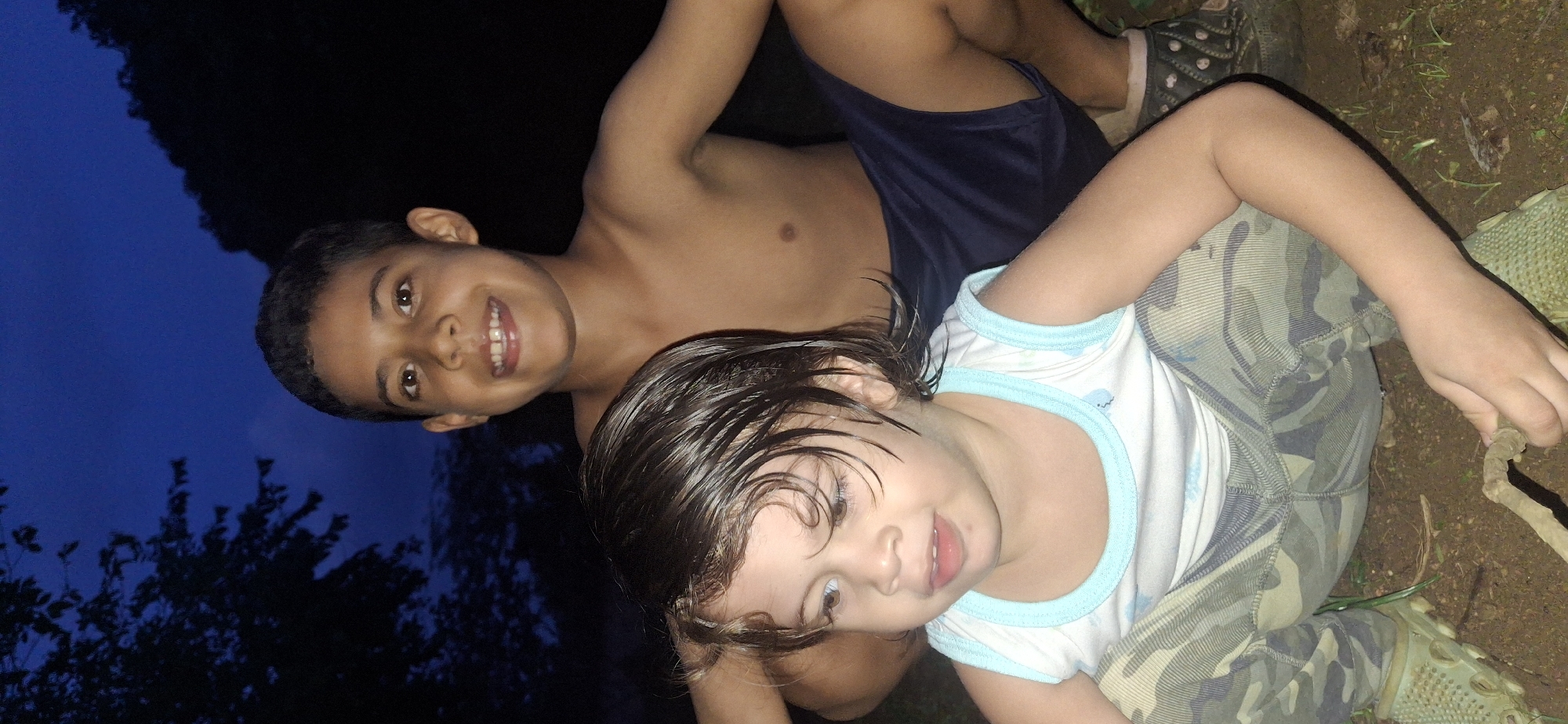 This media file shows a close-up, low-angle shot of two young children in an outdoor setting at twilight. The child in the foreground, on the right, has wet, dark hair and is wearing a white tank top with light blue trim and camouflage pants. Their expression is curious, with wide eyes and an open mouth. They are holding a thin stick or twig.
Behind and to the left of this child is another child, appearing to be slightly older. This child is shirtless and wearing dark blue shorts. They are smiling broadly, showing their teeth. Their head is tilted upwards, and they are looking towards the camera.
The background features a dark, deep blue sky, suggesting it is evening or twilight. Silhouetted trees are visible against the sky on the left side of the frame. The ground appears to be dirt with some scattered leaves and grass. The overall scene has a natural, informal feel, possibly indicating a backyard or wooded area. The children seem relaxed and engaged in playful activity, with smiles suggesting a happy mood. The image is a bit tilted, adding a dynamic perspective to the composition. No visible text is present in the media file.