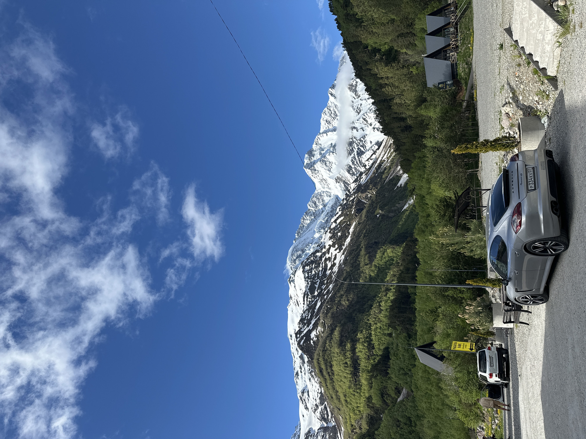 In Terskol, Russia, a silver Porsche Panamera is parked in a gravel lot, facing away from the viewer. To its left, a white SUV with its driver's side door open is parked. In the background, a dense forest of green trees covers the lower slopes of snow-capped mountains, with a clear blue sky overhead. Several modern A-frame cabins are nestled among the trees on the right side of the image. A yellow sign with Russian text is visible near the white SUV.