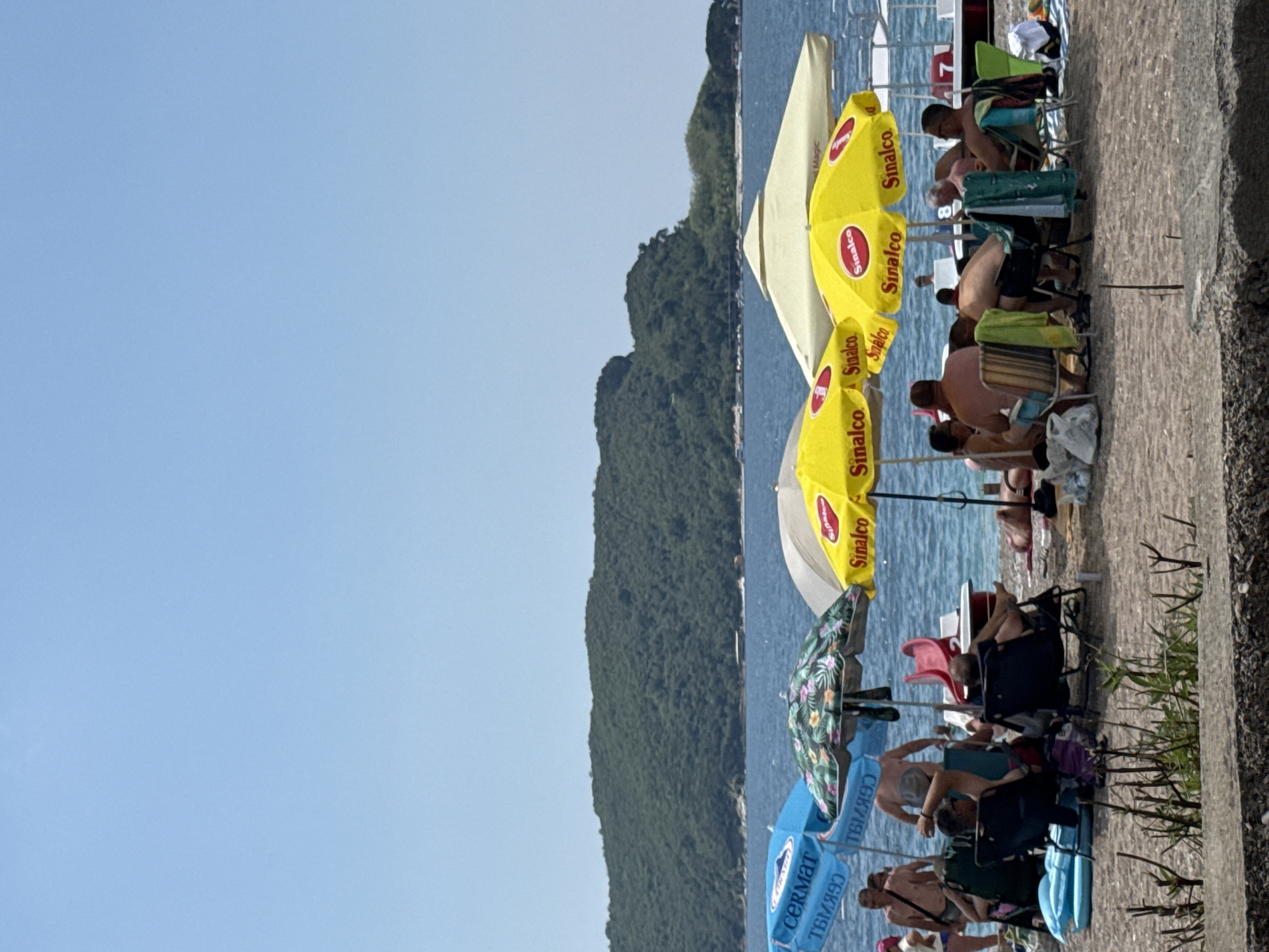 The image depicts a crowded beach scene in Boreti, Montenegro, under a clear blue sky. Several beach umbrellas, including a floral patterned one and yellow "Sinalco" branded umbrellas, provide shade for beachgoers. People are seated in beach chairs, some are lying on towels, and others are standing. In the background, the blue sea is visible, with a green, tree-covered island in the distance. A portion of a boat or watercraft with the number '7' is also visible on the right side of the frame. The ground appears to be a mix of sand and a paved area in the foreground.
