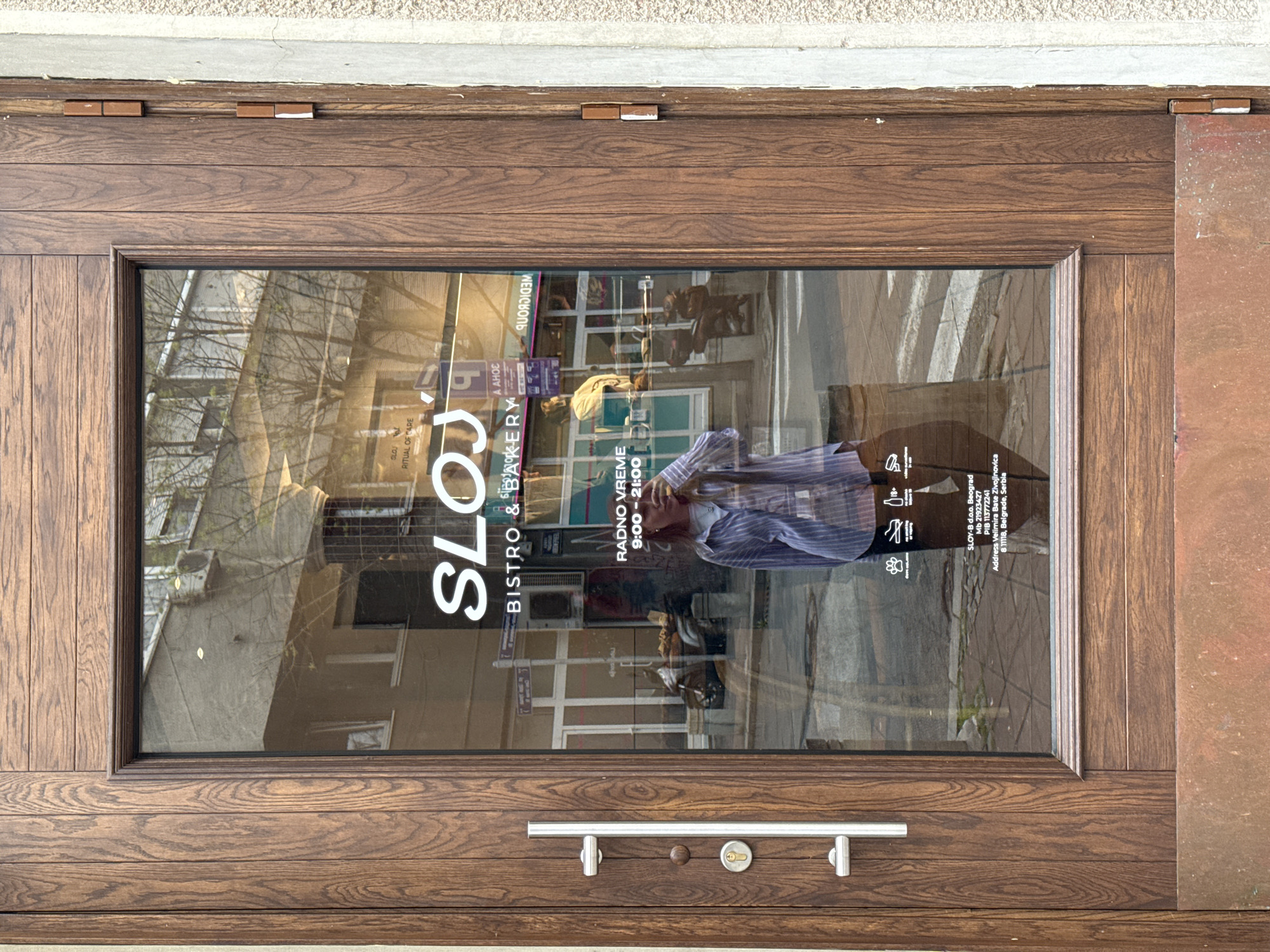 The image shows the front door of "SLOJ Bistro & Bakery" in Belgrade, Serbia. The door is made of wood with a glass window displaying the business name, "SLOJ BISTRO & BAKERY," opening hours "RADNO VREME 9:00-21:00," and company information: "SLOY-B d.o.o. Beograd, Mb 21923427, PIB 113772241, Address Velimira Bate Zivojinovica 8 11118, Belgrade, Serbia." The reflection on the glass shows a person wearing a striped shirt, buildings, and a street scene. The door has a silver handle.