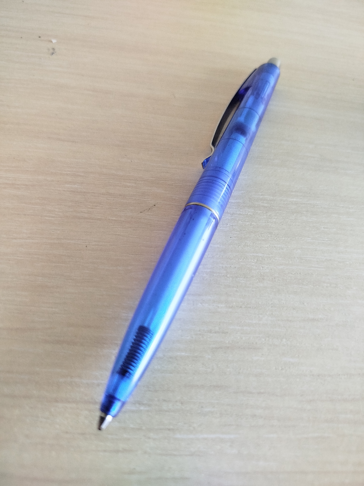 A blue translucent pen with a silver tip and clip is lying diagonally on a light-colored wooden surface. The pen is angled from the top left to the bottom right of the frame. The wooden surface has a visible grain pattern running horizontally. There are a few small dark specks and a small white smudge on the wooden surface.