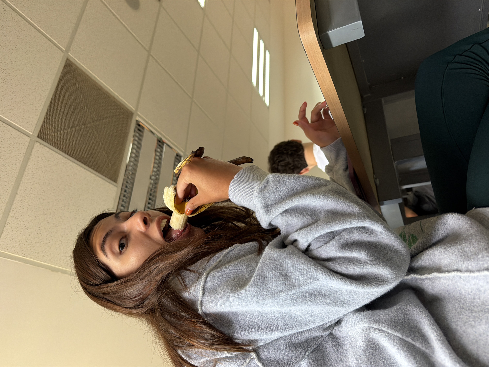 The image, taken in San Andrés Cholula, Mexico, shows an indoor scene focused on a young woman eating a banana, surrounded by other individuals and an institutional setting.
In the foreground, a young woman with long, dark brown hair, wearing a grey hooded sweatshirt, is captured mid-action. She is looking directly at the camera with her mouth wide open, in the process of biting into a partially peeled banana which she holds in her hands. Her expression appears playful or surprised.
Behind her, to the right, the back of another person's head with short, curly brown hair is visible, alongside their hands, suggesting they are seated or engaged in an activity. Further to the right and slightly in the background, a person's leg and hip, dressed in dark green athletic wear, are seen next to a desk or table made of wood and grey metal components.
The background consists of a common indoor ceiling with white acoustic drop tiles, embedded rectangular fluorescent light panels providing illumination, and a large square ventilation grate. This environment suggests a classroom, office, or similar public/educational building. There is no visible text in the image. The lighting is artificial, so the specific time of day is not evident.