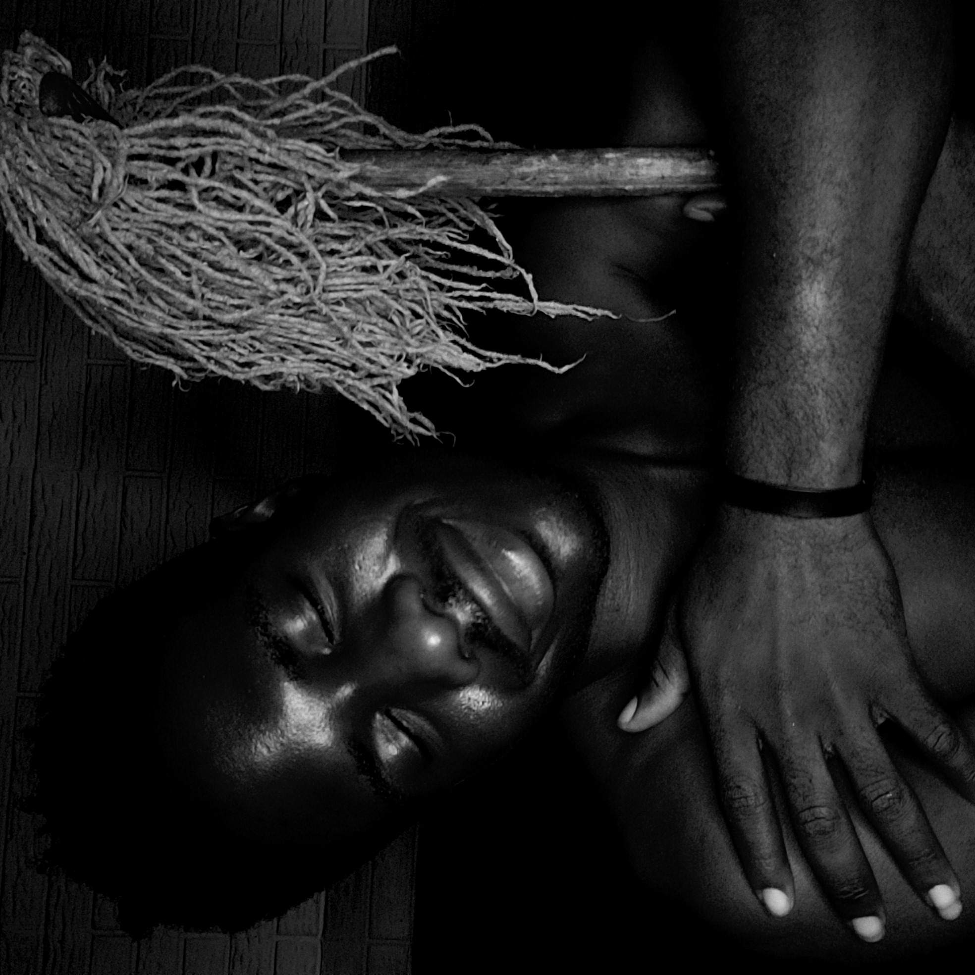 A black and white photograph depicts a dark-skinned man lying on his back, eyes closed, and lips slightly parted. His right arm is bent across his body with his hand resting on his torso. He wears a simple wristband on his right wrist. A bundle of rope or braided material sits above his head, supported by a wooden stick that appears to rest on the side of his face or neck. The background shows a textured surface resembling brick or stone. There is no indication from this image that it is from the city of Narayi, Nigeria.
