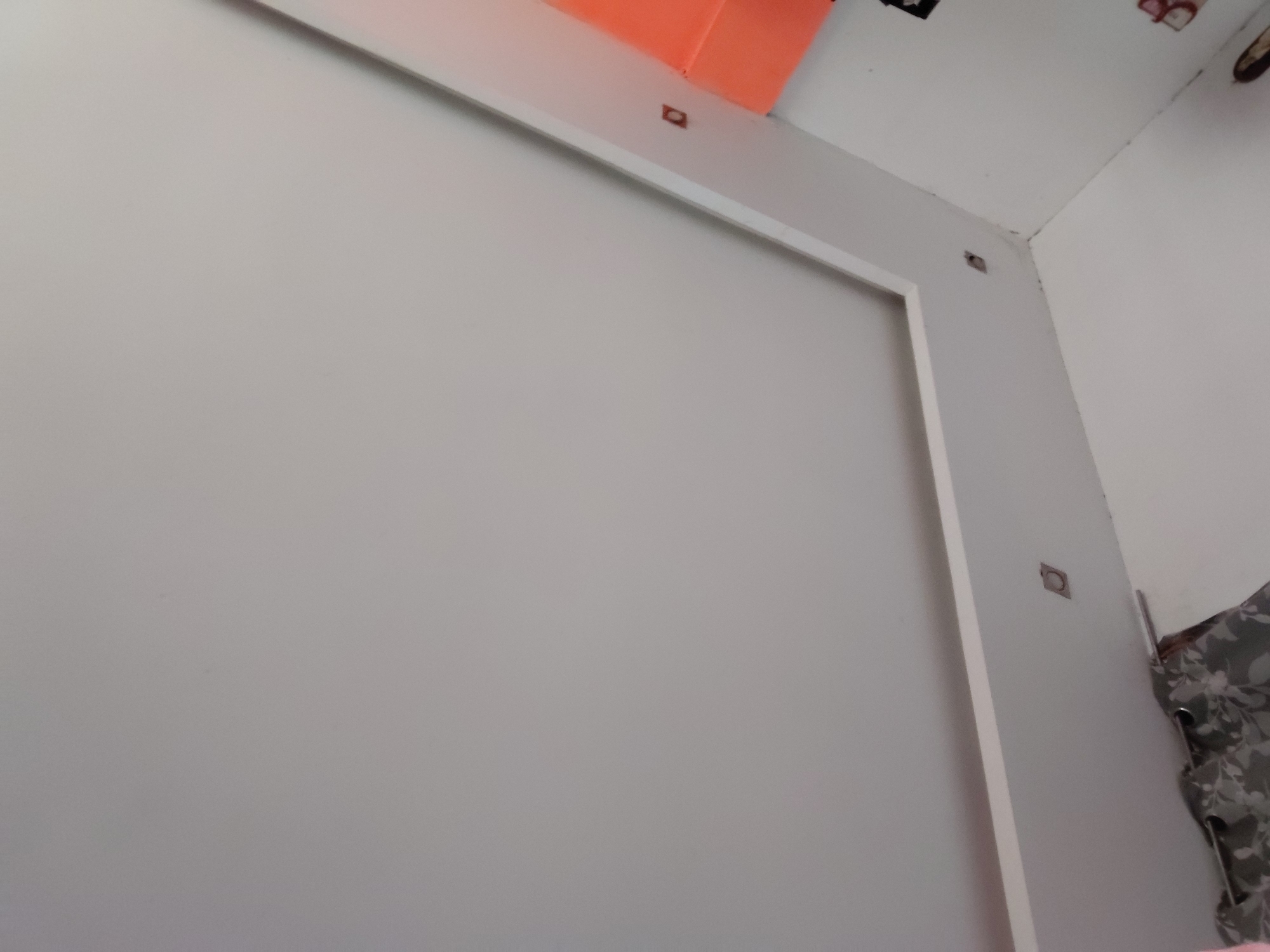The image captures an indoor scene, likely a corner of a room, focusing on a large, plain, light-colored wall or panel framed by white trim. The central subject is a smooth, off-white or light grey surface, appearing to be a wall, a large door, or a built-in panel. This surface is bordered by a clean white frame or trim.
Along the upper horizontal section of the white trim, several small, reddish-brown, square-shaped objects are visible, seemingly embedded into the trim. Another similar object is located on the vertical trim towards the upper right corner. In the top-left portion of the image, an orange object is partially seen, extending downwards from above the white trim.
The upper right section of the image shows a white ceiling and the corner where two white walls meet. Some minor dark smudges or marks are present along the ceiling line. To the far right, a grey curtain with a white floral or leafy pattern is partially visible, indicating a window or a draped area in the room.
No people are present, and no specific activity is taking place. The scene is static, illuminated by what appears to be ambient indoor light. There are no discernible location cues within the image itself to confirm the Santa Ana, Venezuela context, nor is any text visible. The overall impression is that of a simple, functional interior space.