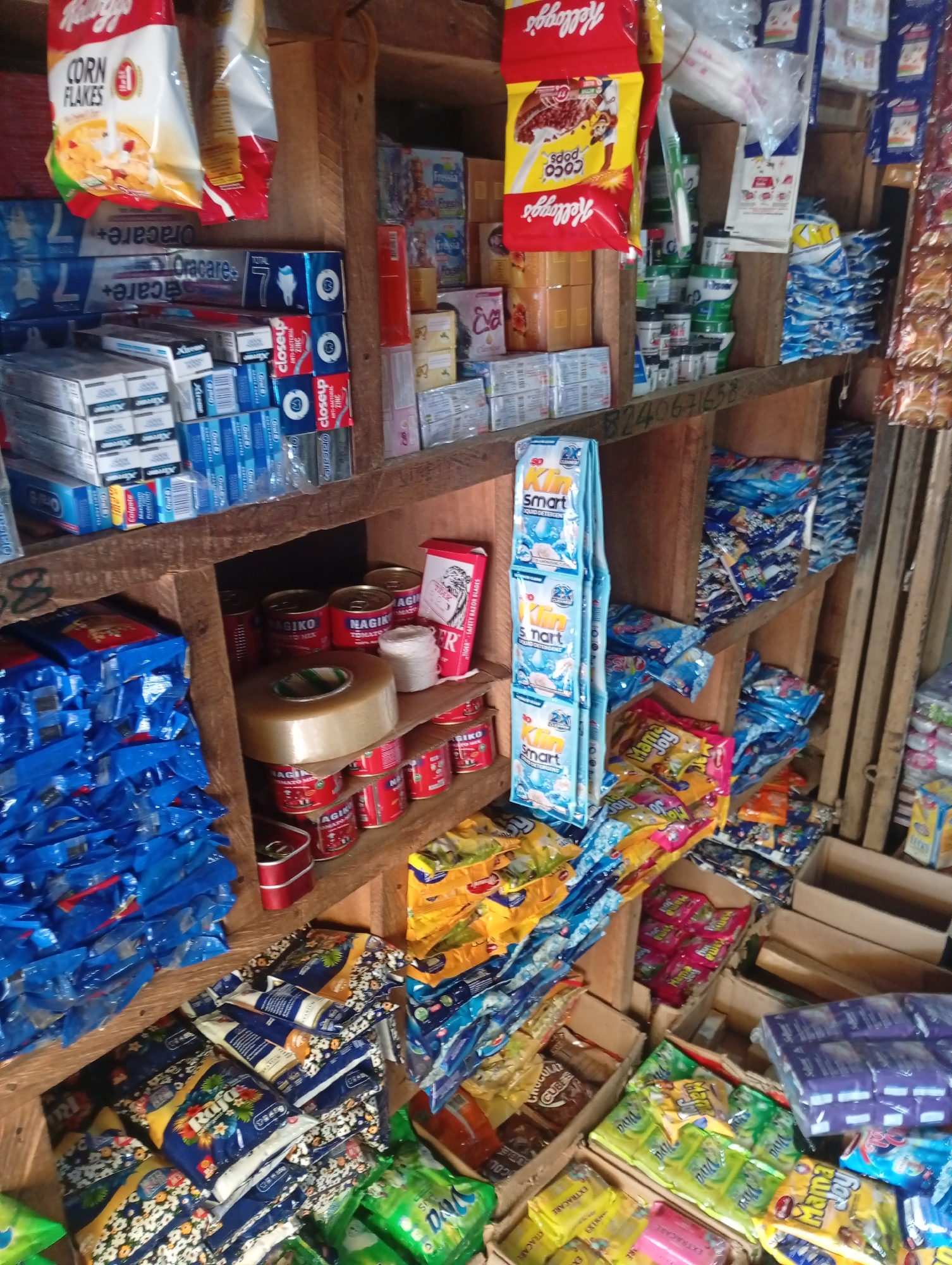 The image depicts a small, cluttered shop interior, likely a market stall, located in Jalingo, Nigeria. The shelves are packed with an assortment of goods, predominantly packaged food items and household products. On the top shelf, red and yellow Kellogg's Corn Flakes and Coco Pops boxes are visible. Below these, rows of toothpaste boxes, including Colgate and Oracare brands, are stacked. To the right, on a higher shelf, are various packaged goods in blue, yellow, and red wrappers, along with some smaller items in clear plastic.
The middle shelf displays an array of canned goods, including "Nagiko Tomato Mix," alongside a large roll of clear tape. There are also packets of what appear to be powdered drinks or detergents hanging vertically, labeled "Klin Smart Liquid Detergent."
The lower shelves are filled with stacks of brightly colored packets, many of which are blue and feature floral or abstract patterns. Some packets are yellow and pink, with branding such as "Mama Joy" visible. There are also boxes containing various smaller packages and some items in clear plastic wrapping. The overall impression is one of a well-stocked, albeit disorganized, local convenience store. The construction of the shelves appears to be made from rough, unfinished wood.