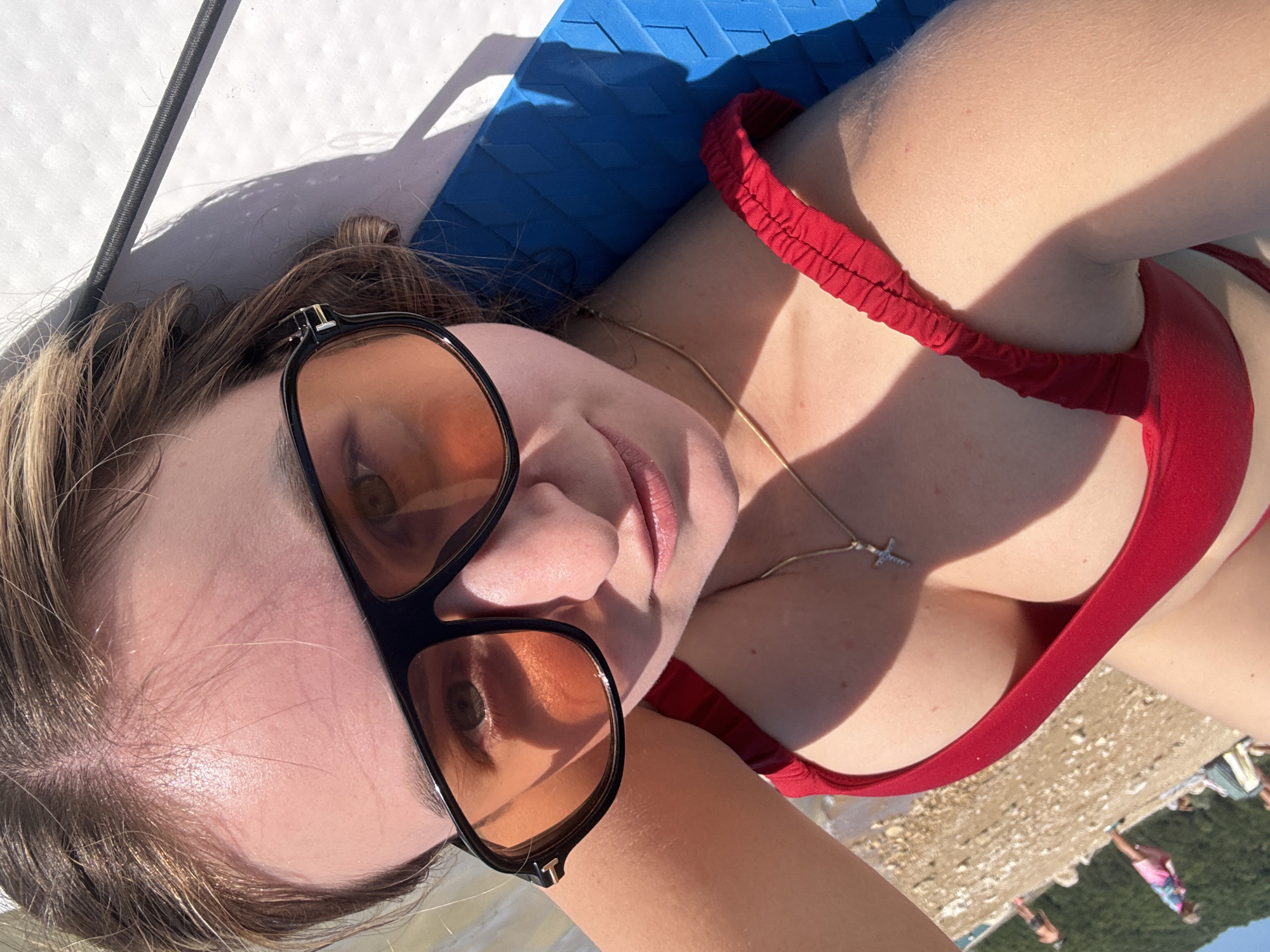 A woman in sunglasses and a red bikini top lies on a blue mat outdoors. Sunlight casts shadows on her face and arm. In the background, a beach with people and water is visible. The image is taken from a high angle, looking down at the woman.