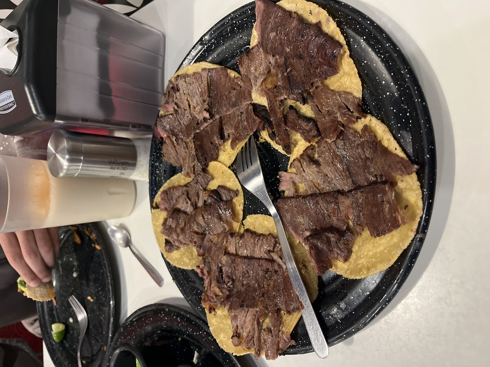 The image depicts a plate of tacos in what appears to be a restaurant setting. The tacos are topped with meat, and a fork rests across them. A black plate with white speckles holds the tacos. To the left, there is a partially visible hand holding a taco, along with a small plate with remnants of food, a spoon, and a lime wedge. A tissue dispenser and a beverage container are also visible. The location is presumed to be in San Bernardino Tlaxcalancingo, Mexico.