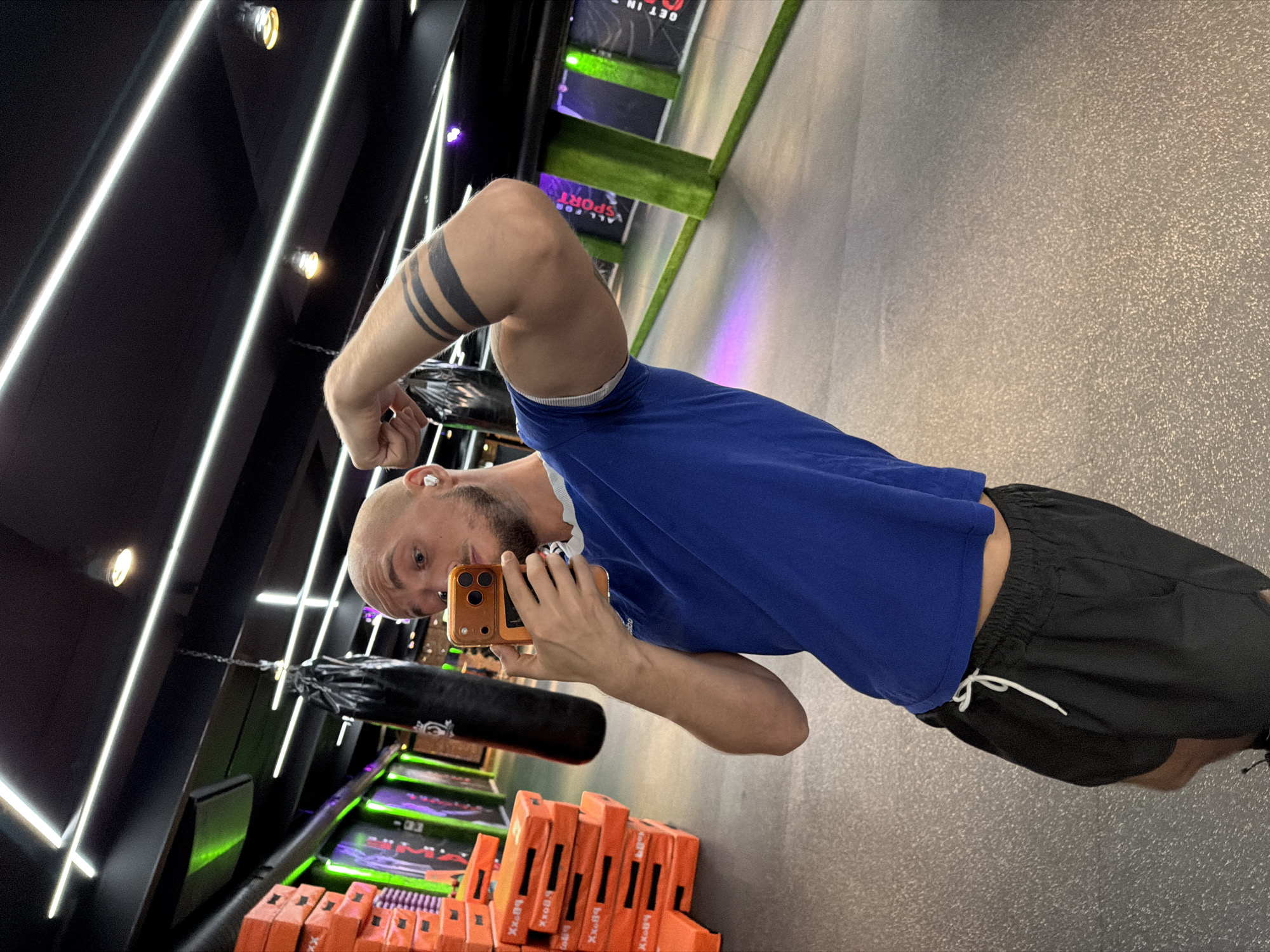 The image depicts a man in a gym, holding a smartphone to take a mirror selfie. He is wearing a blue t-shirt and black shorts, and has tattoos on his arm. The gym appears to be modern, with neon lighting and punching bags. The location is identified as Sofia, Bulgaria.