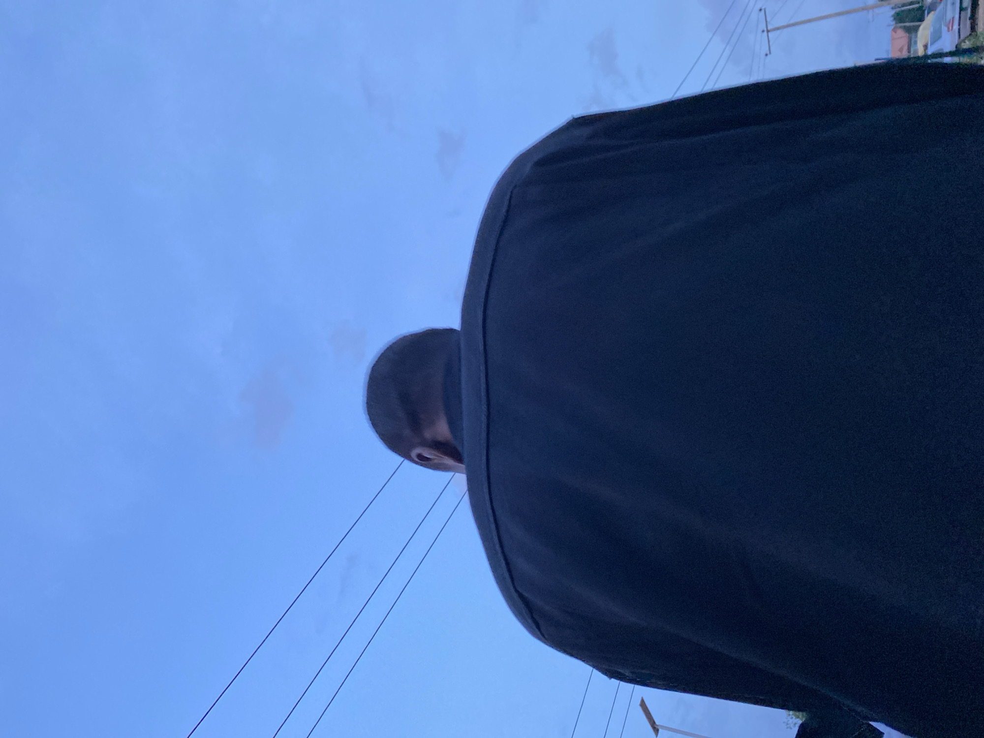 The image shows the back of a person wearing a dark jacket, viewed from a low angle against a cloudy blue sky. Several power lines are visible horizontally across the sky. In the background, a street light and a utility pole can be seen, along with some buildings and a parked car, suggesting an urban or semi-urban environment. The lighting indicates it is likely dusk or dawn. The scene appears to be outdoors in Bukuru, Nigeria, based on the context provided.