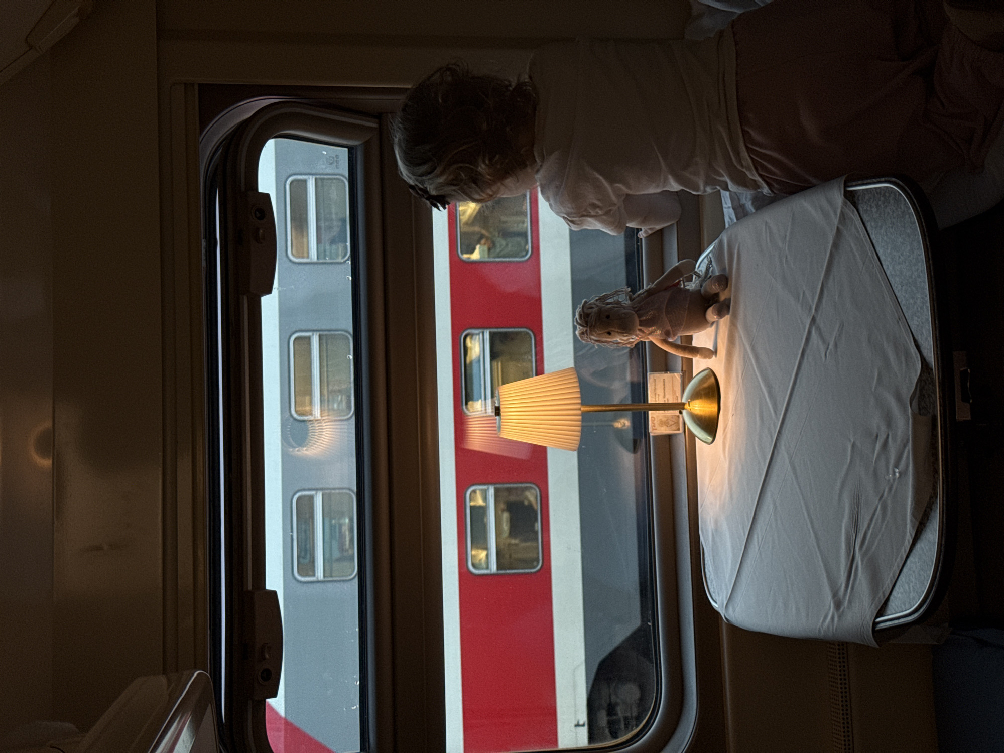 A child is looking out of a train window. Next to them on the table is a lamp and a doll. The train is next to another train car. The train is likely in Russia, but cannot be confirmed.