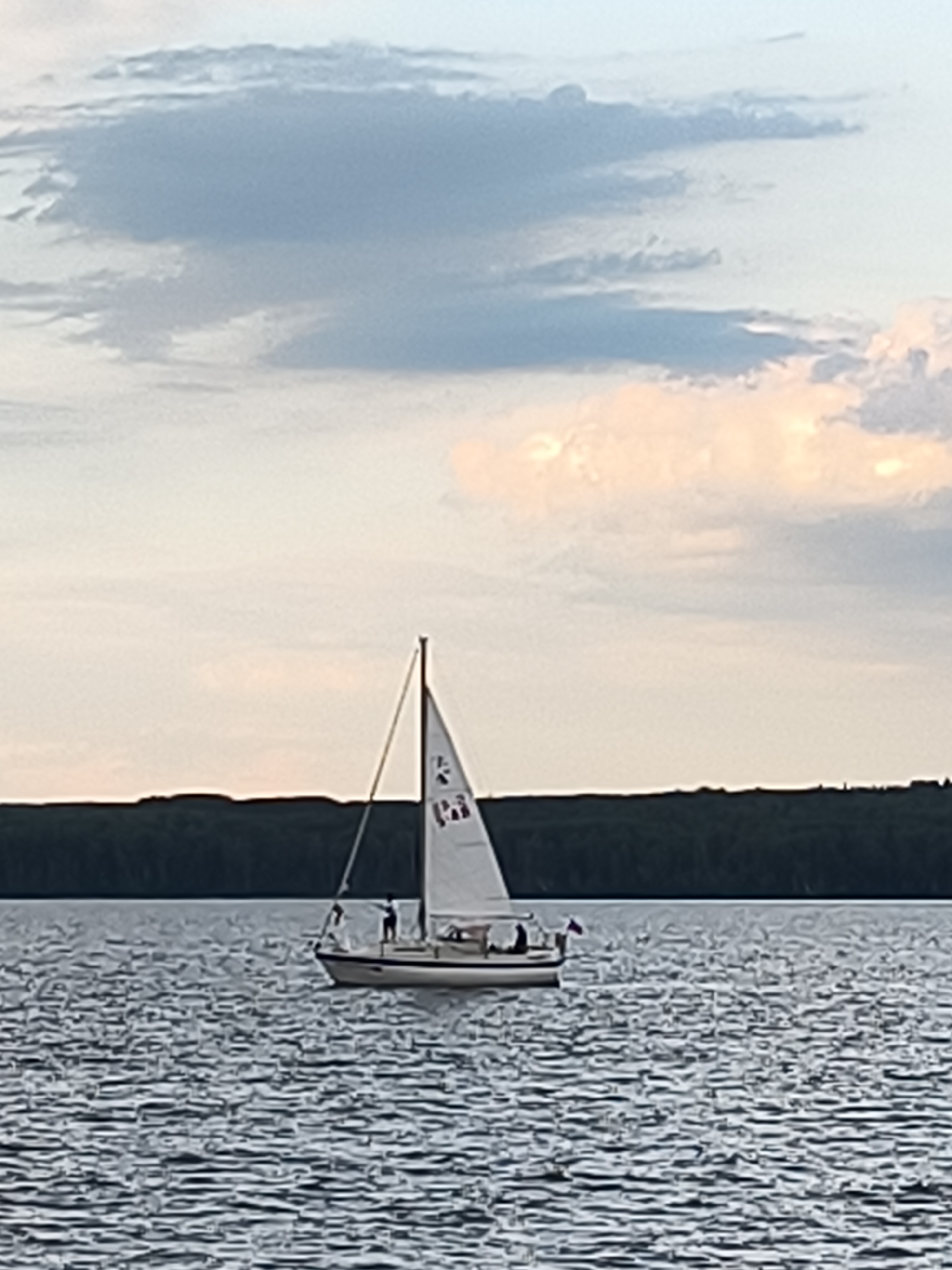 A sailboat with a white sail and a dark hull is sailing on a rippling body of water. Two people are visible on the deck of the sailboat. A dark, tree-lined horizon stretches across the background, separating the water from a sky filled with clouds. The sky has areas of blue and patches of soft, warm-toned clouds, suggesting either sunrise or sunset. The image is captured from a slightly elevated perspective, looking towards the sailboat. The location is not specified as Chaikovsky, Russia.