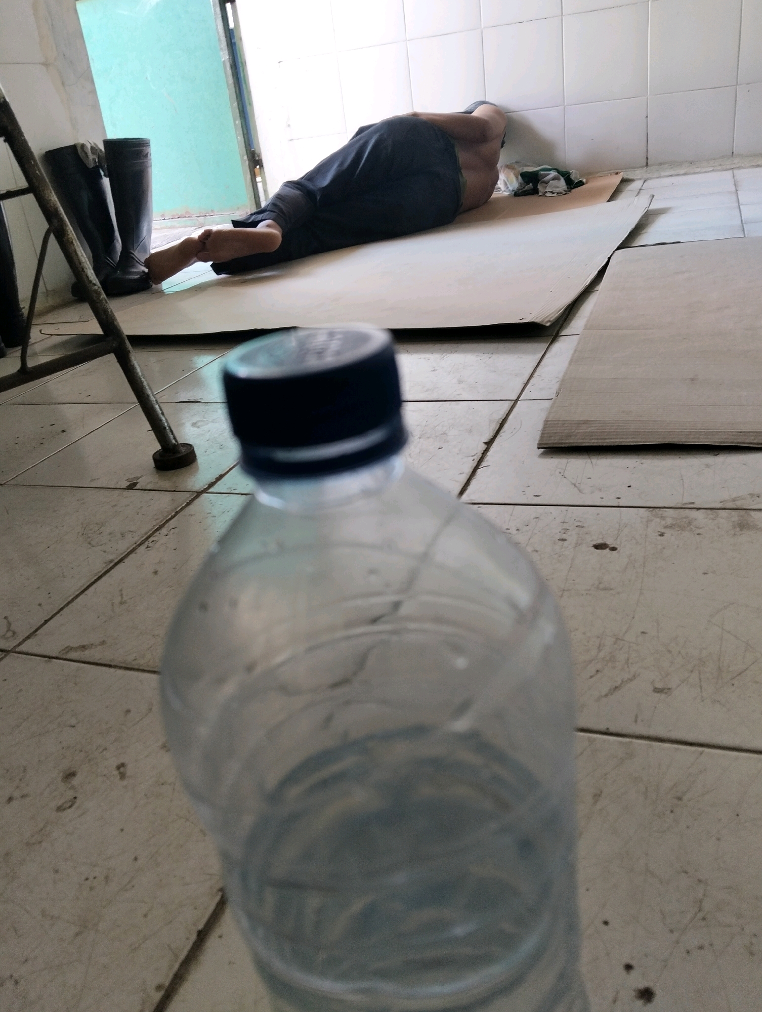 A person is sleeping on a piece of cardboard on the floor in what appears to be an indoor, tiled area, possibly a warehouse or abandoned building. The person is lying on their stomach with their head turned towards a white tiled wall. They are wearing dark pants and their torso is bare. In front of the person are two black rubber boots. To the left of the boots, a metal stand with a circular base is visible. In the foreground, out of focus, is a clear plastic water bottle with a black cap. The background shows a turquoise-colored wall or door and more white tiles. The overall scene suggests a temporary or makeshift sleeping arrangement.