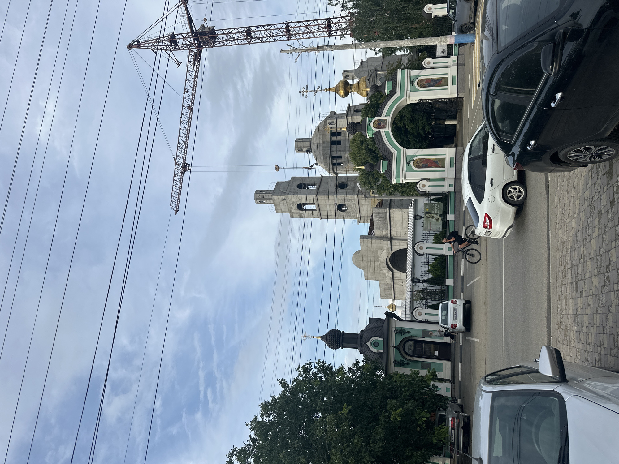 The image shows a street scene in Armavir, Russia, featuring a large church complex under construction, a construction crane, and several vehicles. The foreground is dominated by parked cars and a cobblestone street. In the middle ground, a white car is parked, and a person is riding a bicycle. Behind them is an entrance gate to the church complex, adorned with green and white details and religious imagery. Further back, the church itself is visible, with a large dome and a bell tower, also appearing to be under construction. A prominent red construction crane looms over the scene. Overhead, multiple power lines are strung across the sky, which is partly cloudy. A mature green tree is situated to the left of the church entrance.