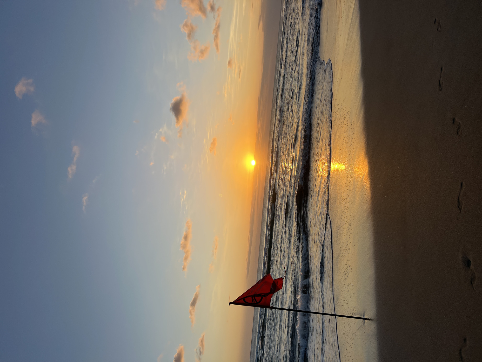 A red flag with a white circular symbol is planted in the sand on a beach in Kuta, Indonesia. The flag is on a dark pole, and it appears to be fluttering in the breeze. The sun is setting over the ocean, casting a warm glow on the water and the sand. Waves are rolling onto the shore, and there are footprints in the sand. The sky is a gradient of orange, yellow, and blue, with a few scattered clouds. The scene is serene and peaceful, with the sound of the waves and the gentle breeze creating a calming atmosphere.