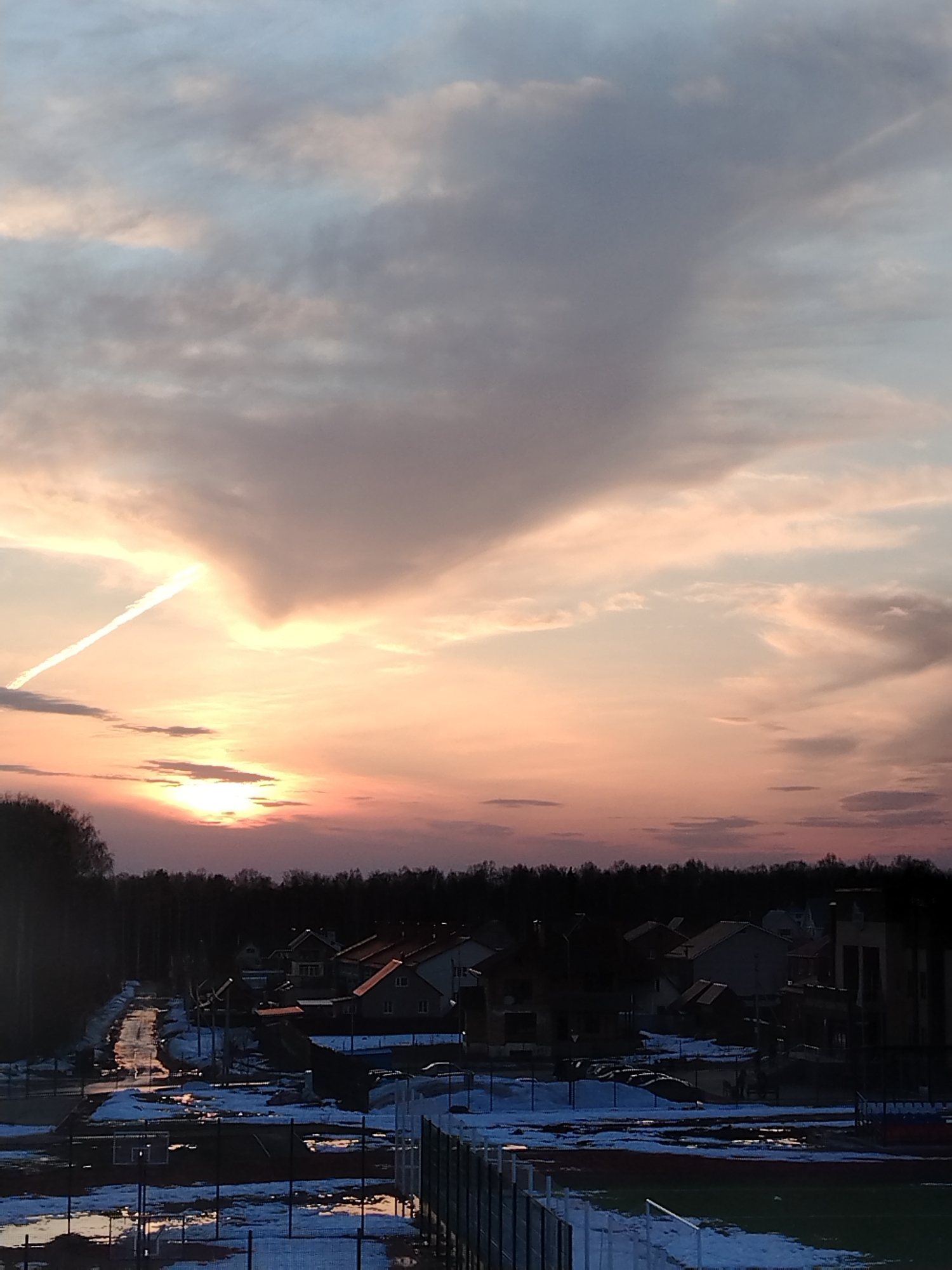 The image shows a sunset over a residential area in Noviy, Russia. The sky is filled with clouds tinged with orange and pink hues from the setting sun. A contrail is visible in the sky. Below, a row of houses is silhouetted against the horizon, with a dark line of trees in the background. In the foreground, a sports field with remnants of snow and puddles of water is visible. A fence borders the field.