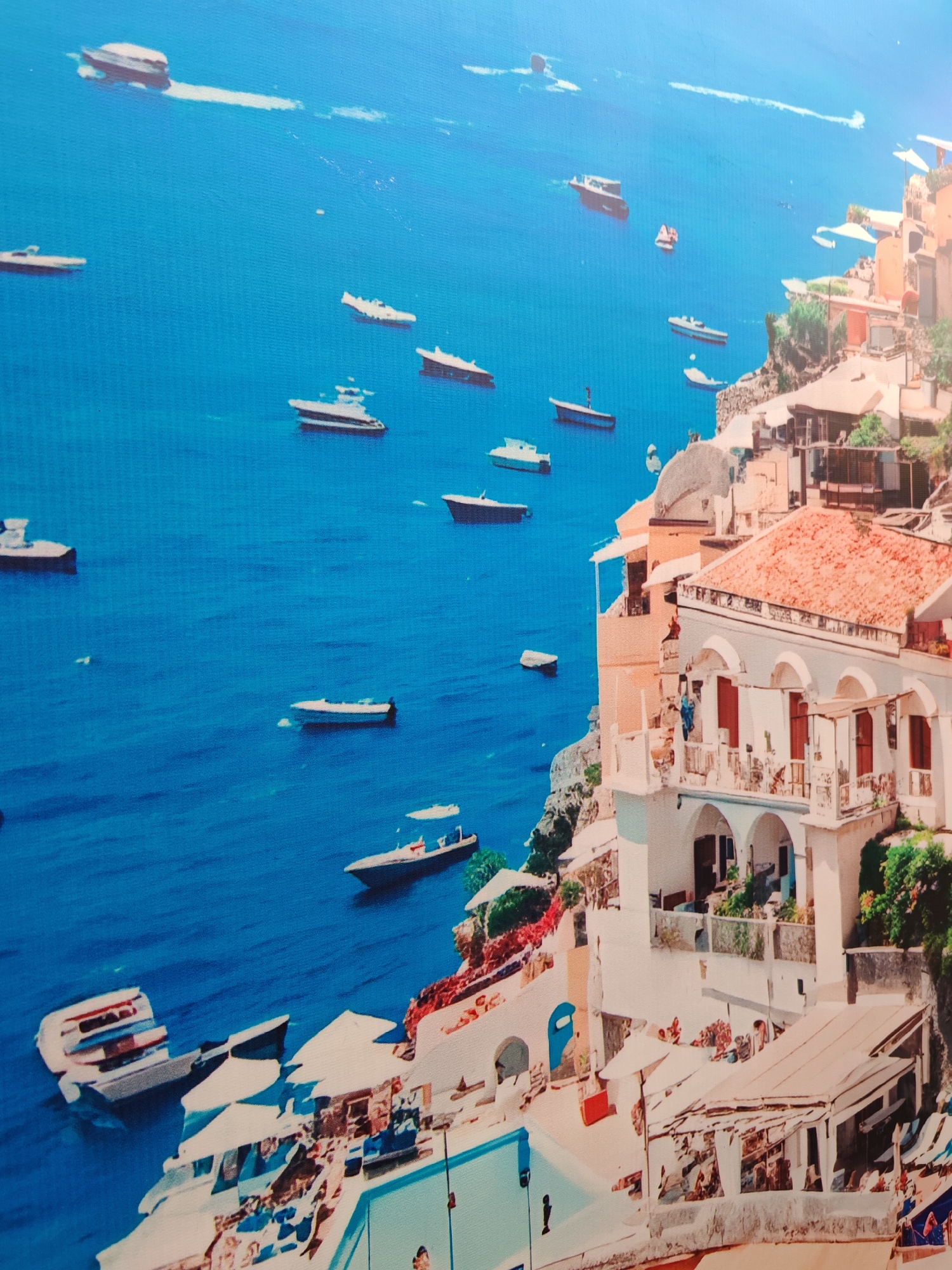 The media file displays a vibrant, picturesque coastal town built into a steep cliffside, overlooking a calm, deep blue sea under bright daylight. Numerous boats and yachts of various sizes, predominantly white, are scattered across the water, some leaving faint white trails.
The town features densely packed, multi-tiered buildings, primarily whitewashed or light-colored, with distinctive terracotta or reddish-brown tiled roofs. Many structures include balconies, arched windows, and outdoor terraces with umbrellas and seating areas, suggesting residential or resort accommodations. A light blue swimming pool is visible in the lower-left foreground, surrounded by more terraced structures and umbrellas. Small patches of green vegetation cling to the cliffs and dot the architectural landscape.
The scene conveys a sense of leisure and tranquility, typical of a popular tourist destination. The architectural style is reminiscent of Mediterranean coastal towns. The image itself appears to be a photograph of a canvas print or a digital image with a noticeable textured overlay, showing a slight weave pattern across its surface. No discernible text is visible within the image.