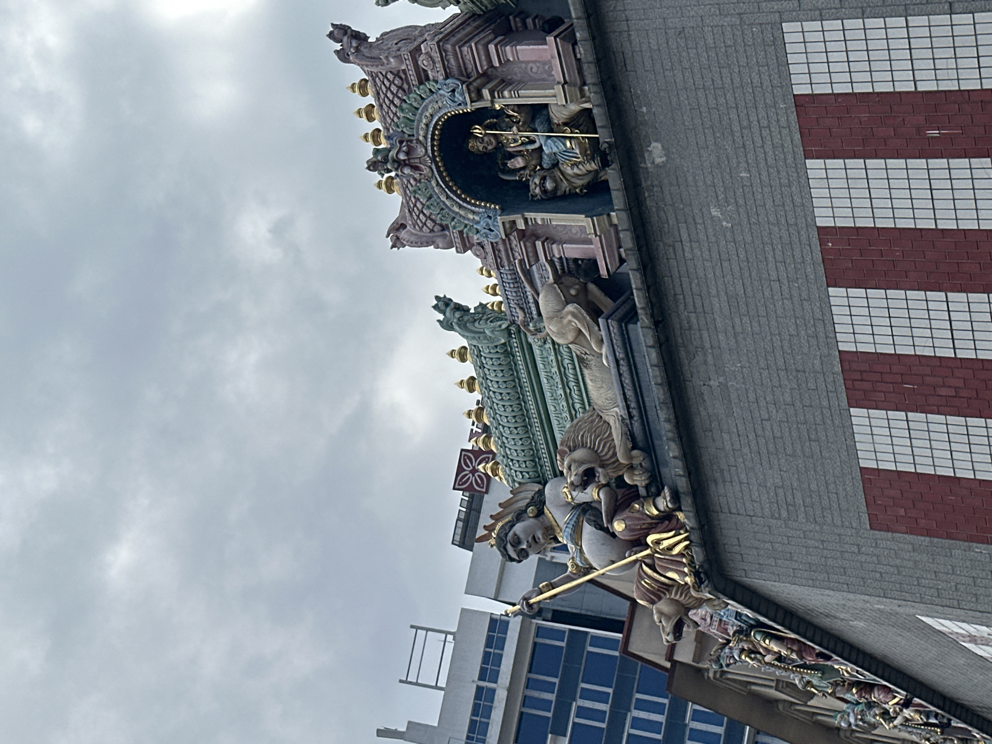 The image shows the exterior of a building, possibly a temple, with intricate sculptures adorning its rooftop. These sculptures depict various figures and animals, including what appears to be a deity sitting with a lion and another figure within an ornate archway. In the background, modern buildings with glass windows are visible, suggesting an urban setting. The sky is overcast with clouds. The building itself is characterized by a gray brick facade and decorative bands of red and white tiles. The overall scene captures a blend of traditional religious art and contemporary architecture in Singapore.