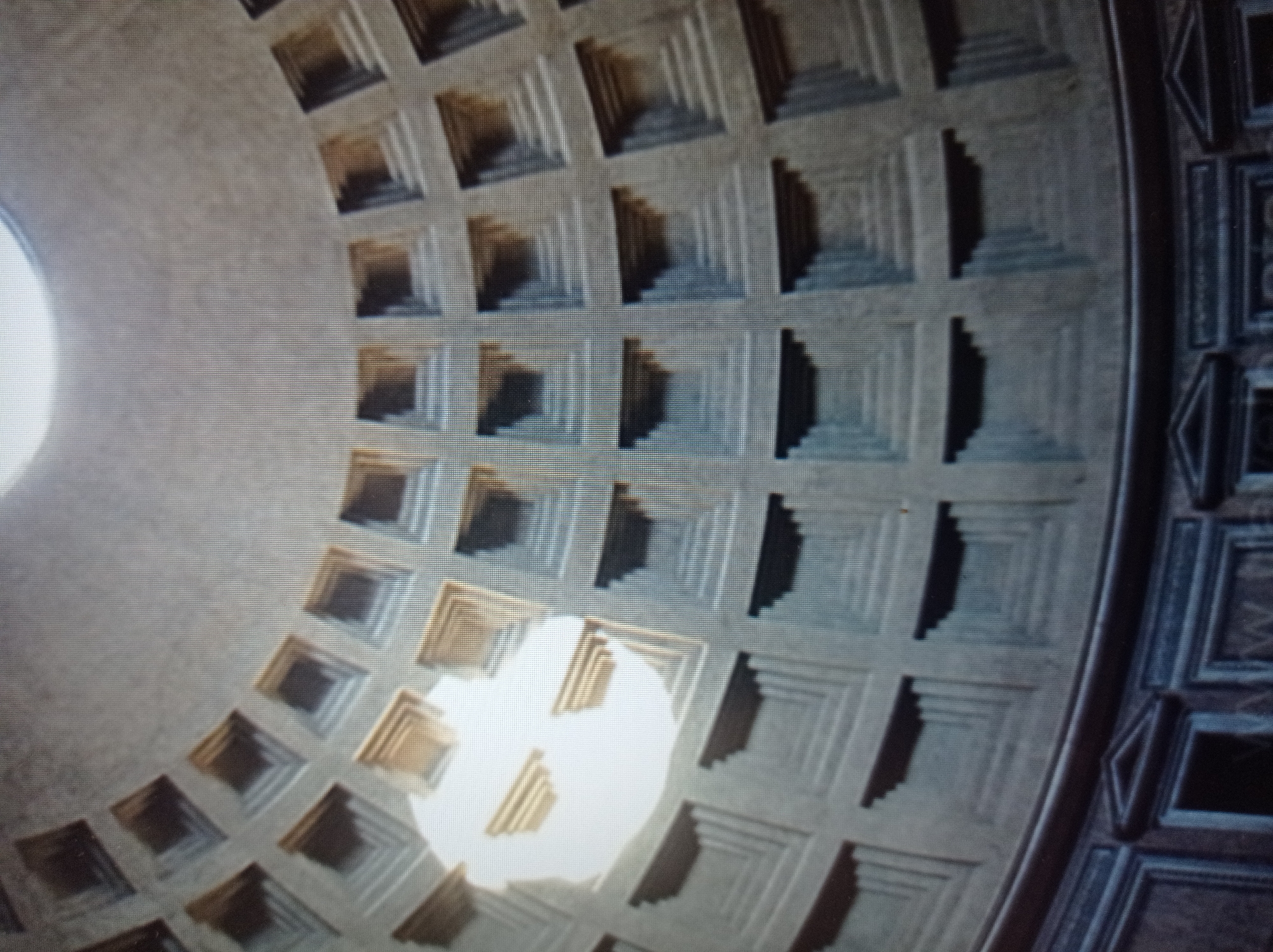 The image shows the interior of a dome, likely the Pantheon in Rome, Italy, due to its characteristic coffered ceiling and oculus. The dome is constructed from concrete and features a circular opening at the apex, allowing natural light to illuminate the interior. The coffered ceiling consists of concentric rings of square or rectangular coffers, each recessed and adorned with a stepped pattern, creating a sense of depth and grandeur. The lower portion of the image reveals the curved walls of the building, which are also decorated with architectural elements. There is a bright light source emanating from the oculus, casting shadows and highlighting the texture of the coffered ceiling. The overall impression is one of immense scale and architectural sophistication. The provided context of Mukachevo, Ukraine, is inconsistent with the architectural style depicted in the image, which is characteristic of ancient Roman architecture.