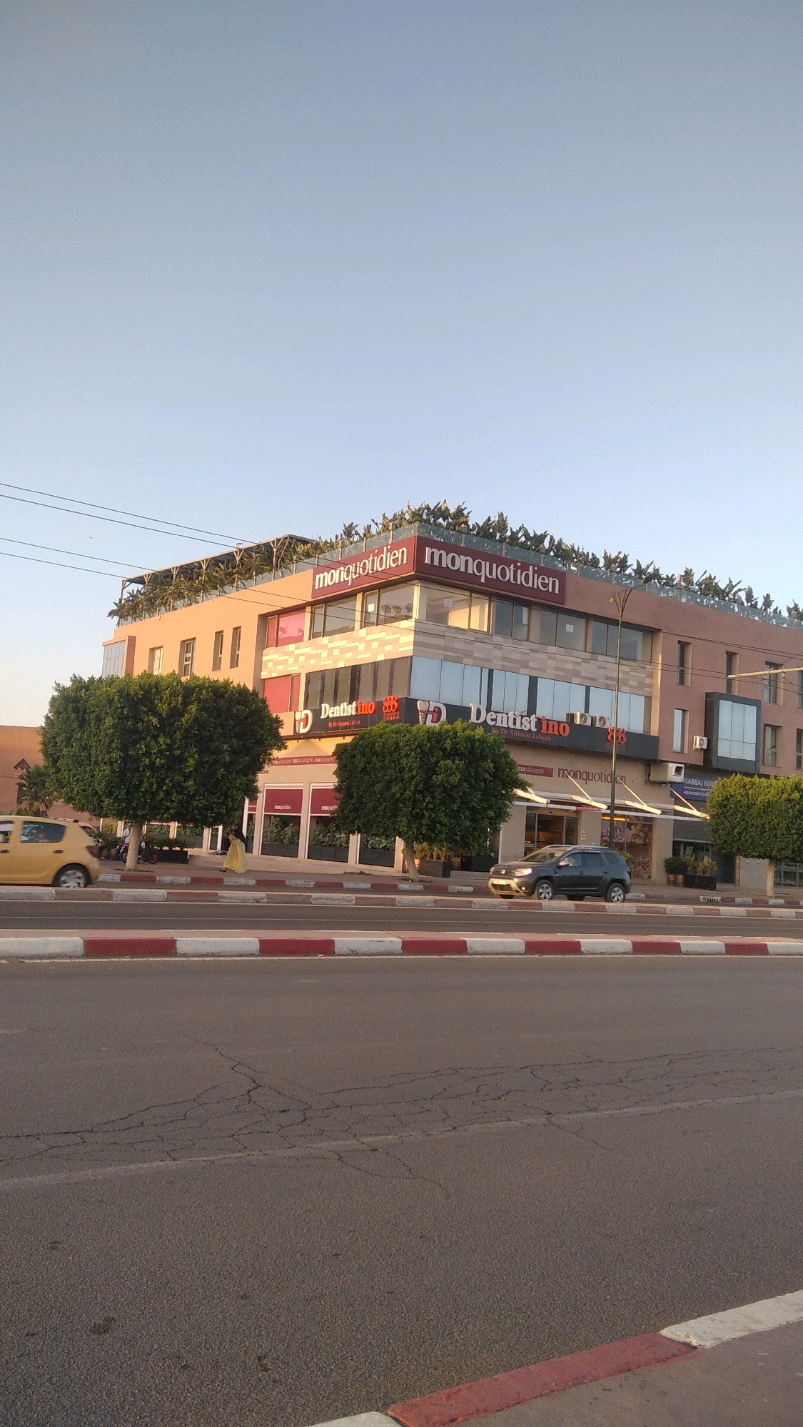 The image shows a modern, multi-story building on a sunny day in Marrakech, Morocco. The building features glass windows and prominent signage, with "monquotidien" and "Dentistino" visible. Two well-maintained, rounded trees stand in front of the building. A yellow car is parked on the left, and a dark-colored car is driving on the right side of the street, which is paved with asphalt and bordered by a curb with red and white markings. The sky is clear and pale blue, suggesting it is late afternoon or early morning.