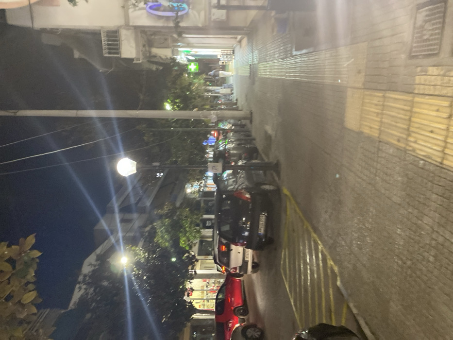 The image captures a nighttime street scene in an urban area, specifically Egaleo, Greece. The perspective looks down a street flanked by buildings, with a focus on artificial lighting and parked vehicles.
The sky is dark, indicating that it is night. Artificial light sources, particularly a bright street lamp in the mid-ground, illuminate the scene, casting strong starburst flares. Buildings line both sides of the street, some with visible air conditioning units. On the left side, several cars are parked bumper-to-bumper, including a black hatchback with a partial Greek license plate ("IYP-14XX" where 'X' denotes unclear digits), a white car, and a red car in the foreground. Yellow painted lines mark sections of the asphalt, likely indicating parking zones. A white sign on a pole is visible among the cars, but its text is illegible.
The right side of the street features a wide sidewalk paved with textured stones, including strips of yellow tactile paving. Further down the street, various storefronts or establishments are visible. A prominent green cross sign, commonly used for pharmacies in Europe, is clearly seen. Another circular blue neon sign is visible further in the background. Trees and foliage are interspersed along the street, contributing to the urban landscape. No discernible people are clearly visible, but the presence of businesses and parked cars suggests a typical, albeit quiet, evening in an urban setting.