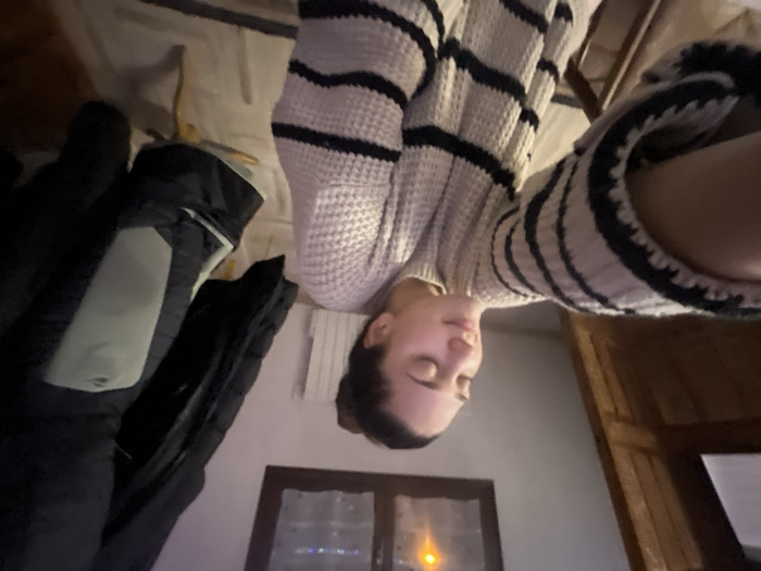 A person wearing a striped sweater is positioned in the center. The room is slightly dark with a window visible in the background. A black jacket with a gray patch hangs on the left. To the right is a wooden cabinet. The photo has a disorienting perspective, with the person and objects rotated, creating an unusual viewpoint.